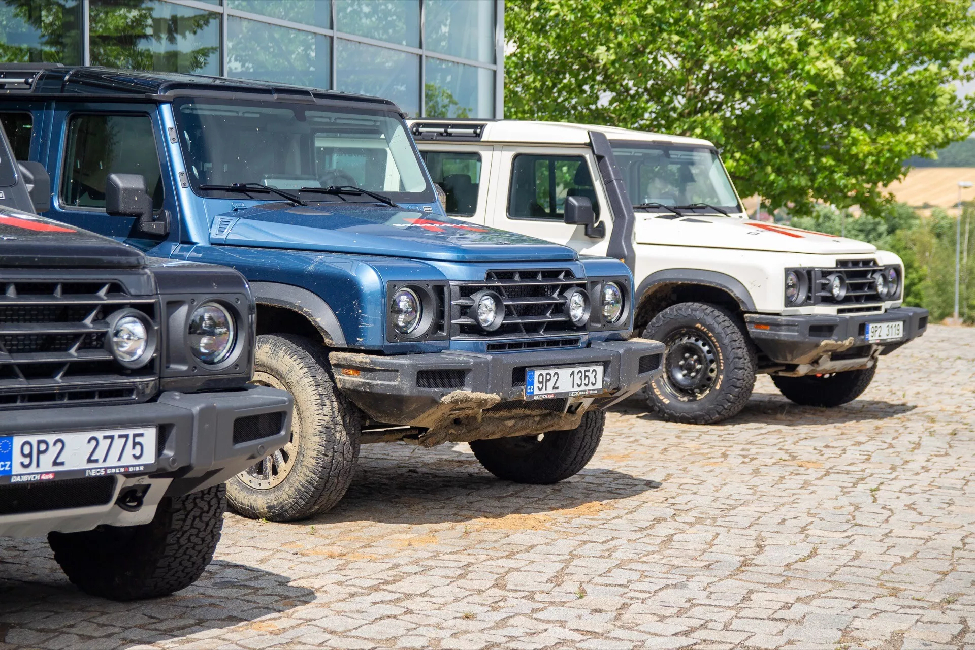 Sestava první generace Ineos Grenadier 2022, zahrnující tři SUV s doplňky pro off-road parkované na parkovišti, prezentující přední mřížku, světlomety a robustní hranatý design
