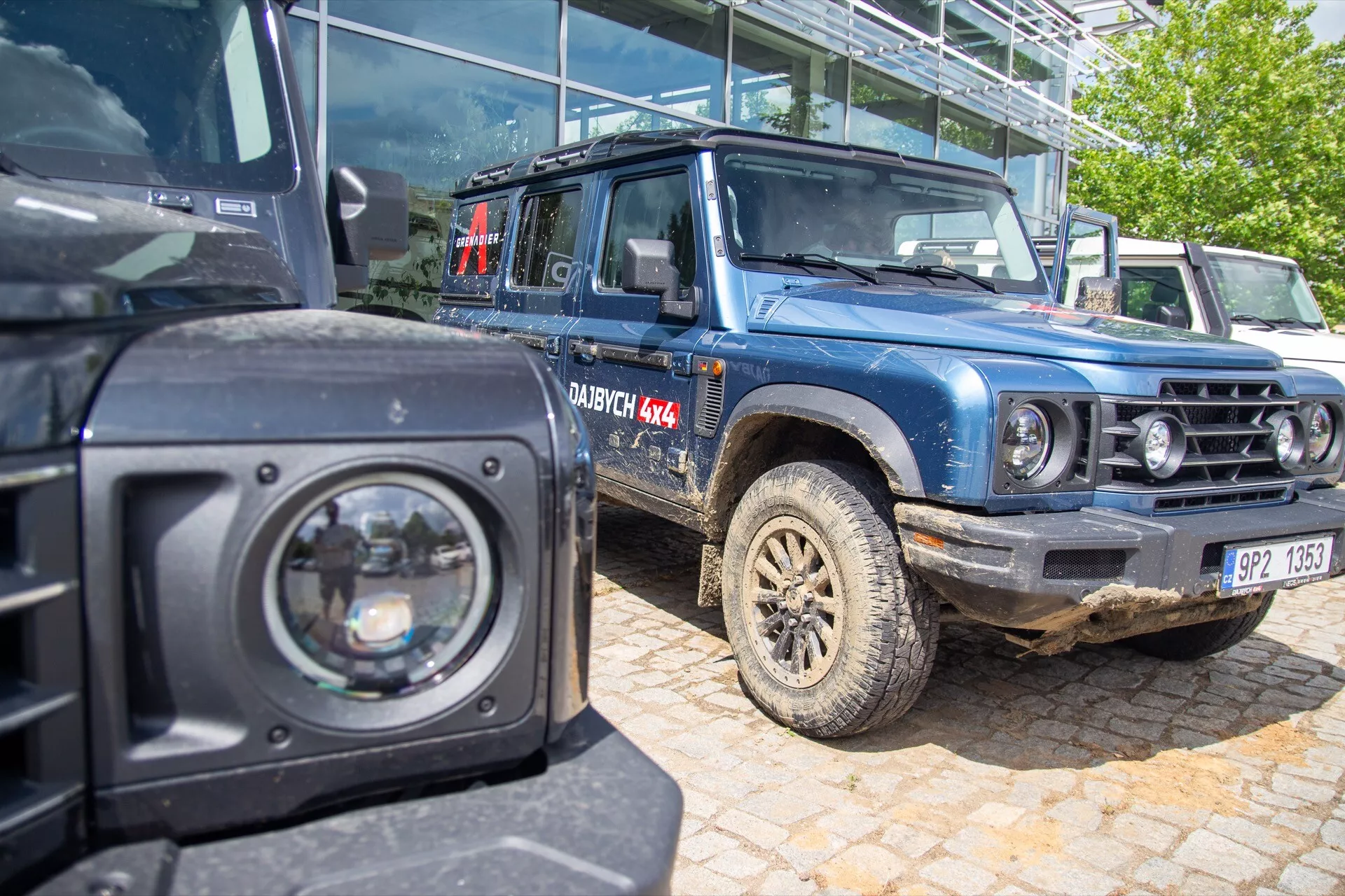 Středně blízký pohled na modrý Ineos Grenadier první generace 2022 se zablácenými pneumatikami a značením DAKAR 4X4, ukazující schopnosti pro terénní jízdu se šnorchlem, reflektory a bezpečnostními reflexními prvky, viděn z čelně-boční perspektivy vedle dalšího černého SUV