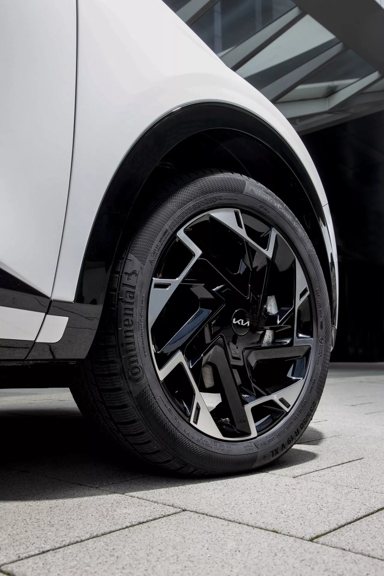 Detailní pohled na kolo Kia Sportage 2022 NQ5 s úhlovým vícepaprskovým designem a pneumatikou Primacy s označením EV, na pozadí bílého bočního karoserie vozidla včetně předního blatníku a dveří řidiče
