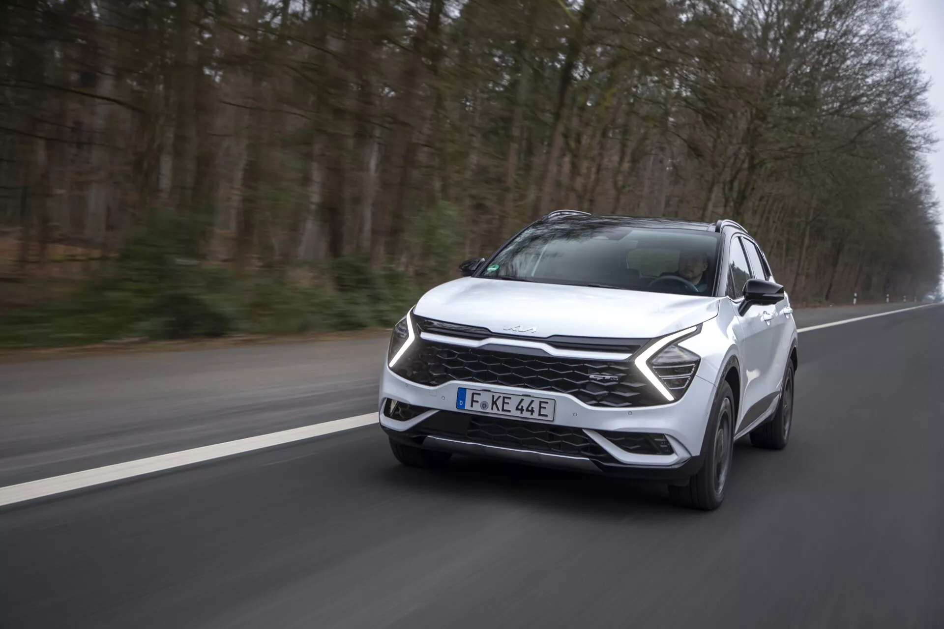 Dynamický pohled z přední strany na Kia Sportage NQ5 rok 2022 v pohybu, předvádějící aerodynamický design a pokročilé technologické funkce, jako jsou LED světlomety a přední senzory ADAS, fotografováno na silnici s provozem vpravo.