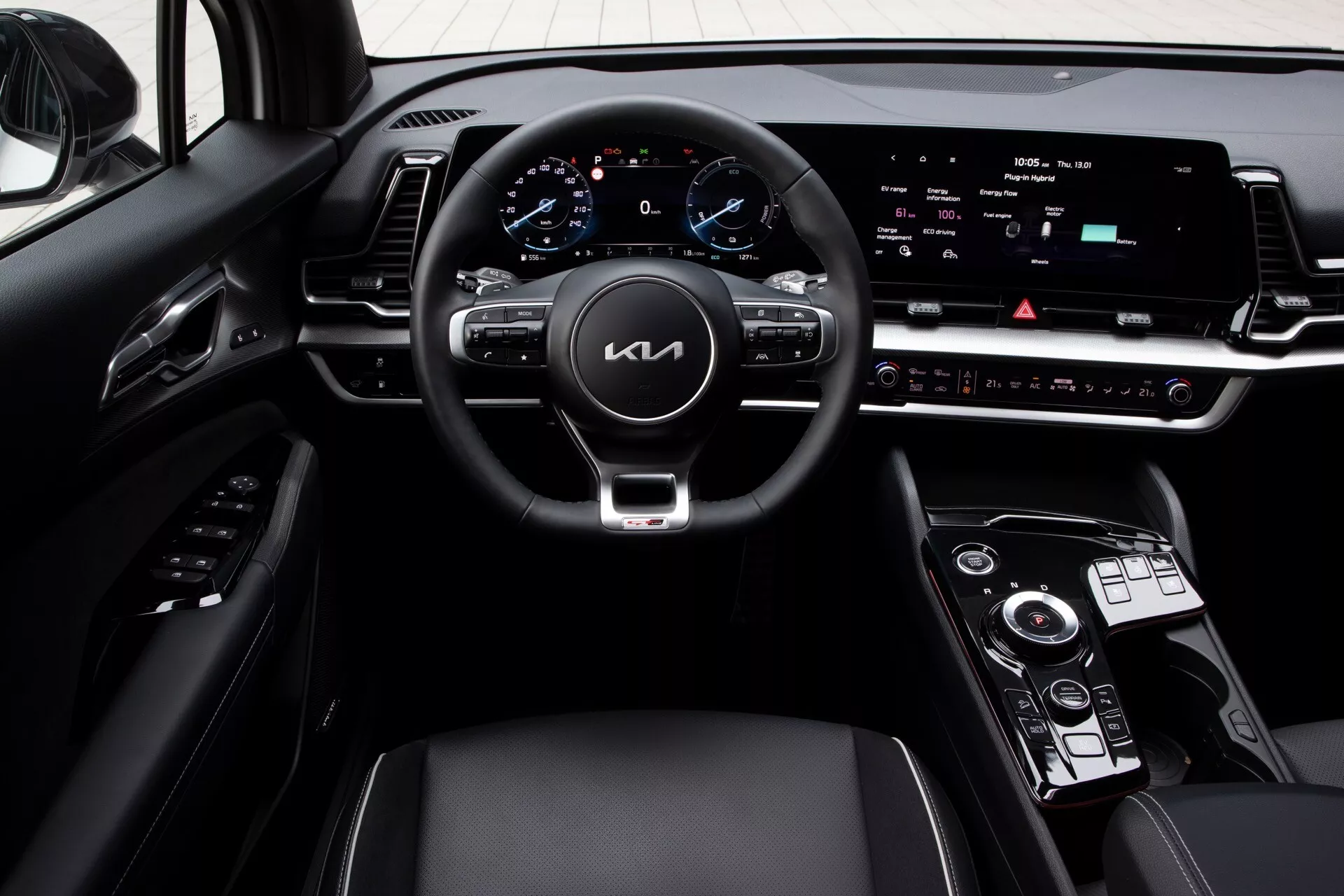 Interiér Kia Sportage (NQ5) 2022 s pohledem na palubní desku řidiče z perspektivy středové konzoly, obsahující tříramenný volant s integrovanými ovladači, digitální přístrojový panel a širokoúhlý centrální informační systém, spolu s otočným ovladačem převodovky a ovládacími prvky klimatizace.
