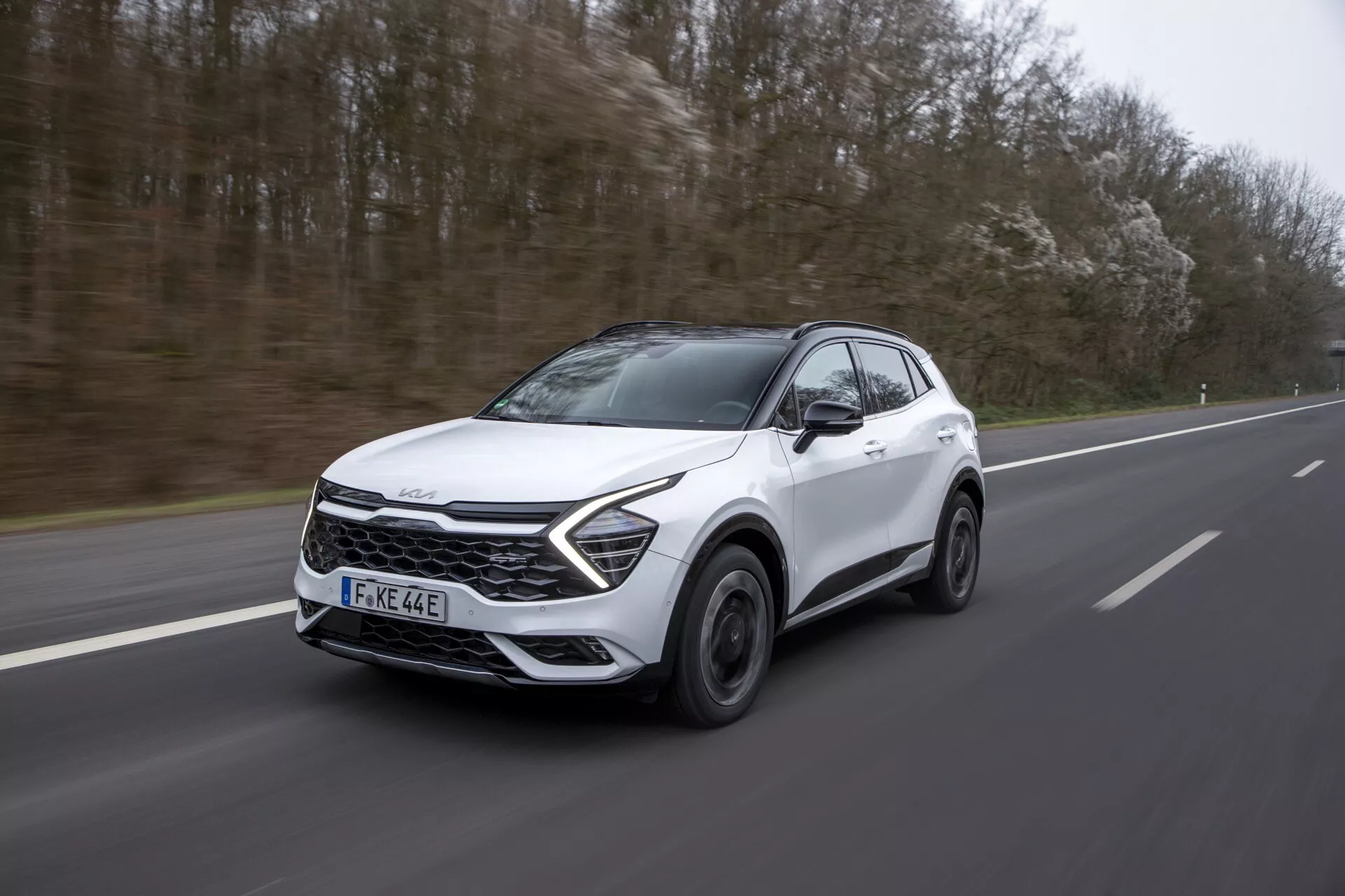Kia Sportage NQ5 2022 v pohybu na dálnici, předvádí svůj aerodynamický bílý exteriér, slitinová kola, tónovaná okna, LED světlomety, moderní design mřížky a dvoubarevný obklad za zataženého osvětlení