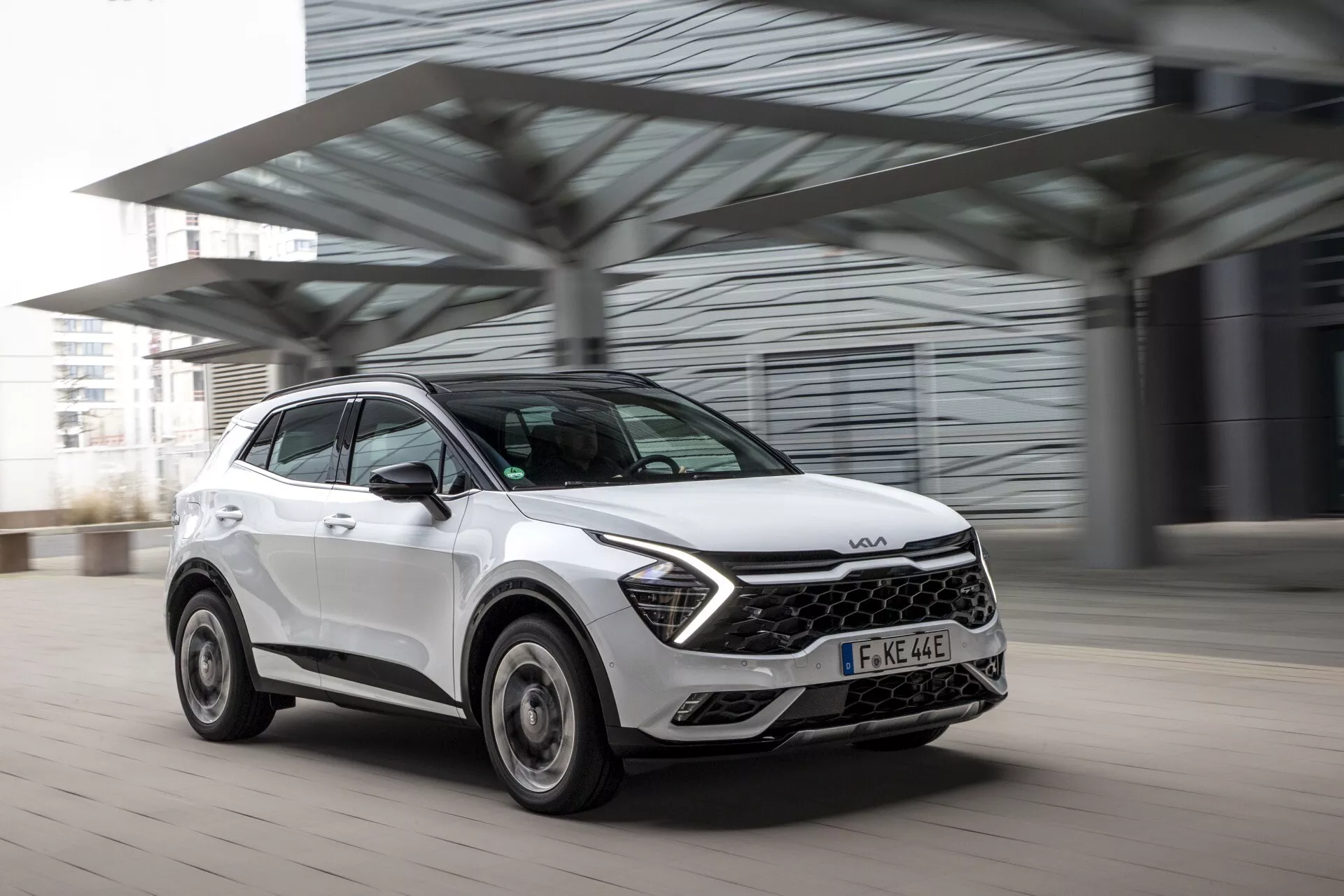 Kia Sportage NQ5 2022 - moderní bílý crossover/SUV v pohybu, zachycen z předního trojčtvrtečního úhlu, který zdůrazňuje přední mřížku, LED světlomety a dynamický design s rozmazaným pozadím naznačujícím pohyb