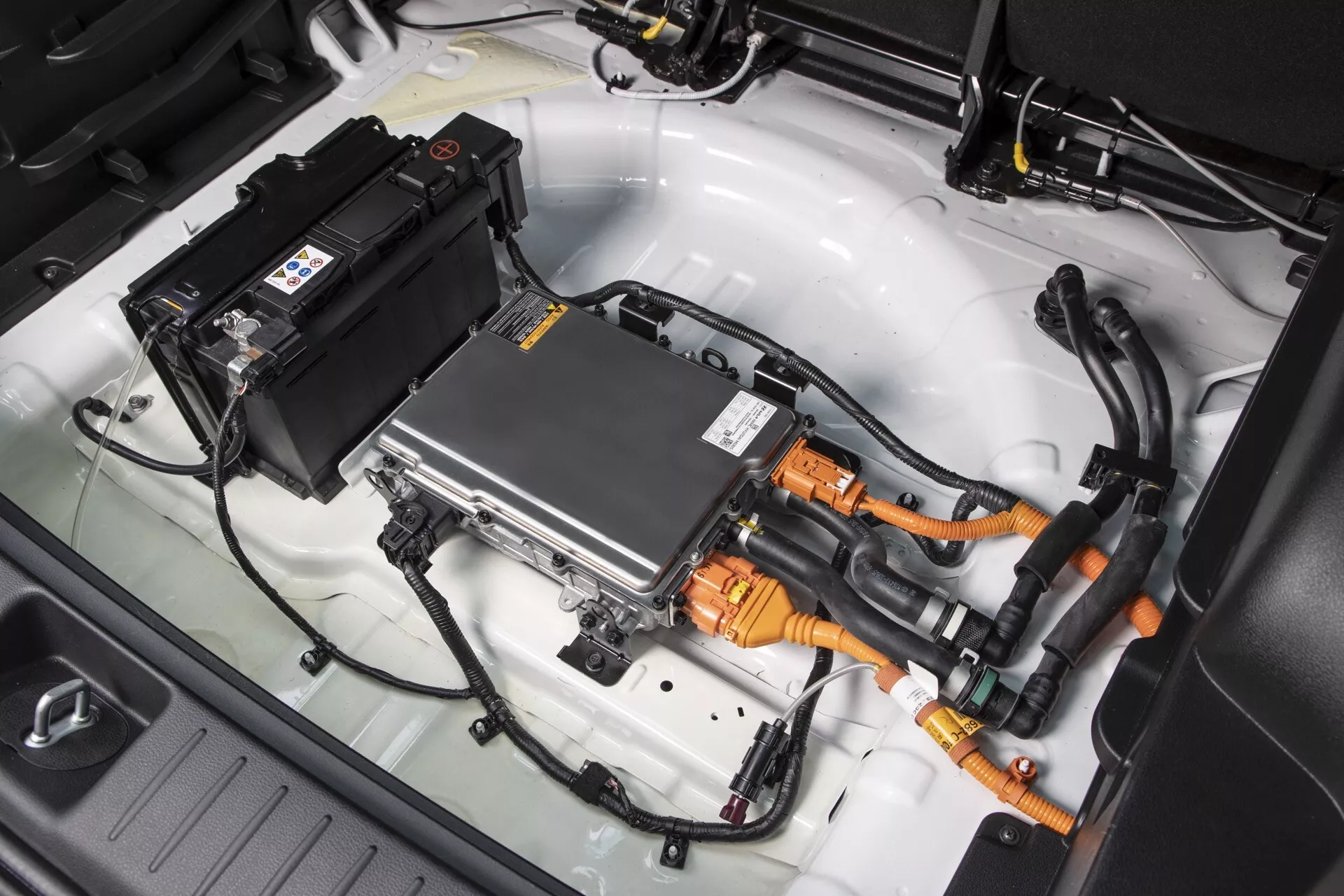 Detailní pohled na komponenty pohonu elektrického nebo hybridního vozu Kia Sportage 2022 NQ5, obsahující velký bateriový blok, vysokonapěťové oranžové kabely, stříbrný invertor s chladiči a integrované karosářské provedení vozidla.