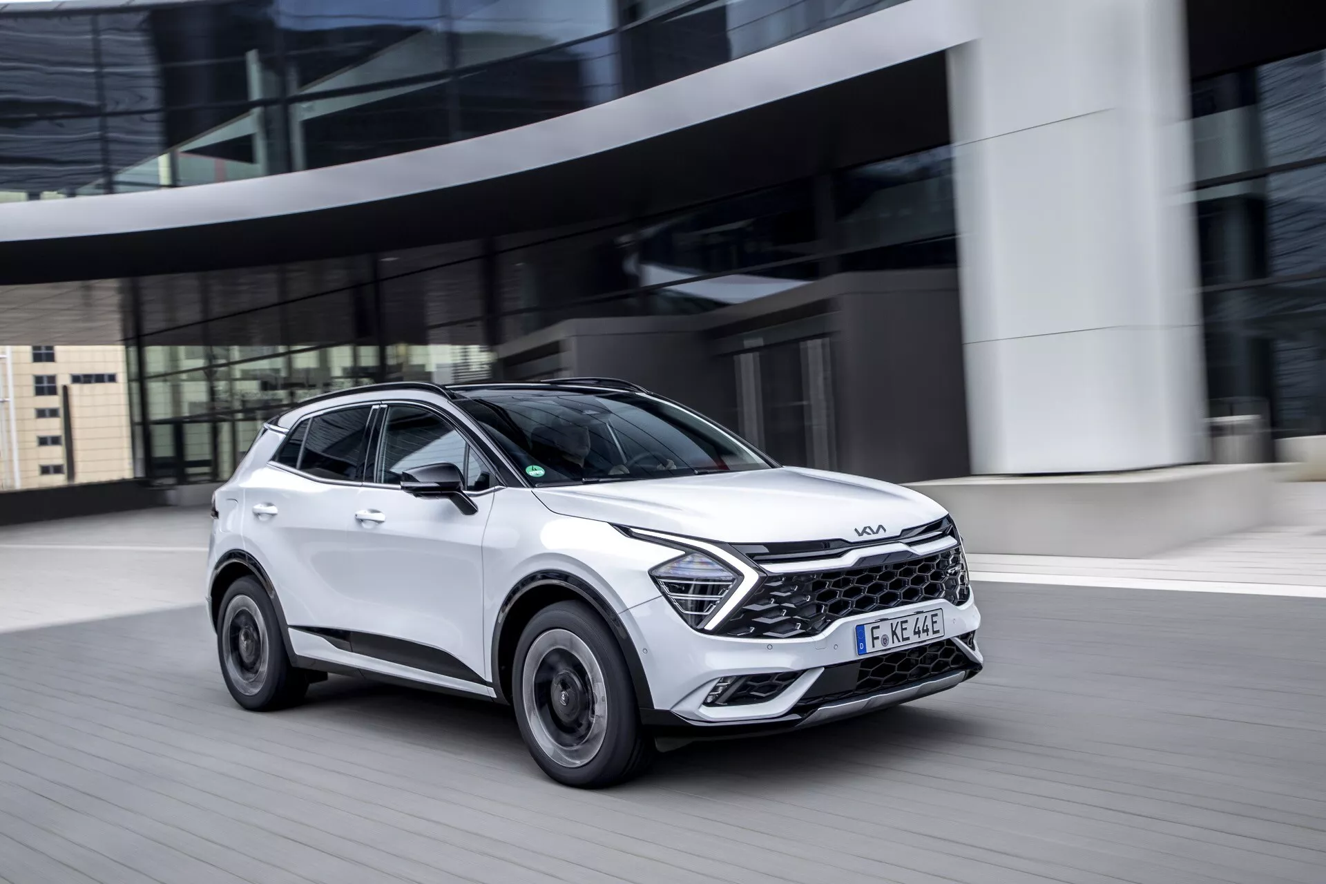 Kia Sportage NQ5 2022 v pohybu, bílé kompaktní SUV s černým lemem a dynamickým designem na městské silnici, předvádějící LED světlomety a sportovní přední mřížku