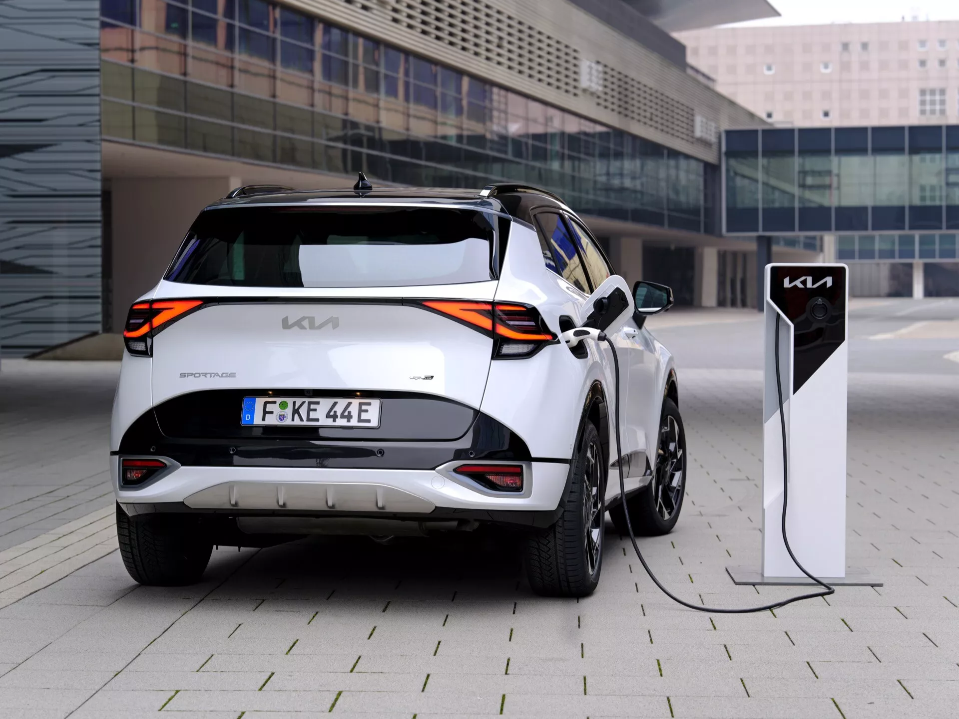 Zadní tříčtvrteční pohled na bílý elektrický vůz Kia e-Niro zaparkovaný a připojený k nabíjecí stanici, prezentující charakteristická zadní světla, znak 'Kia', evropskou poznávací značku a aerodynamická litá kola