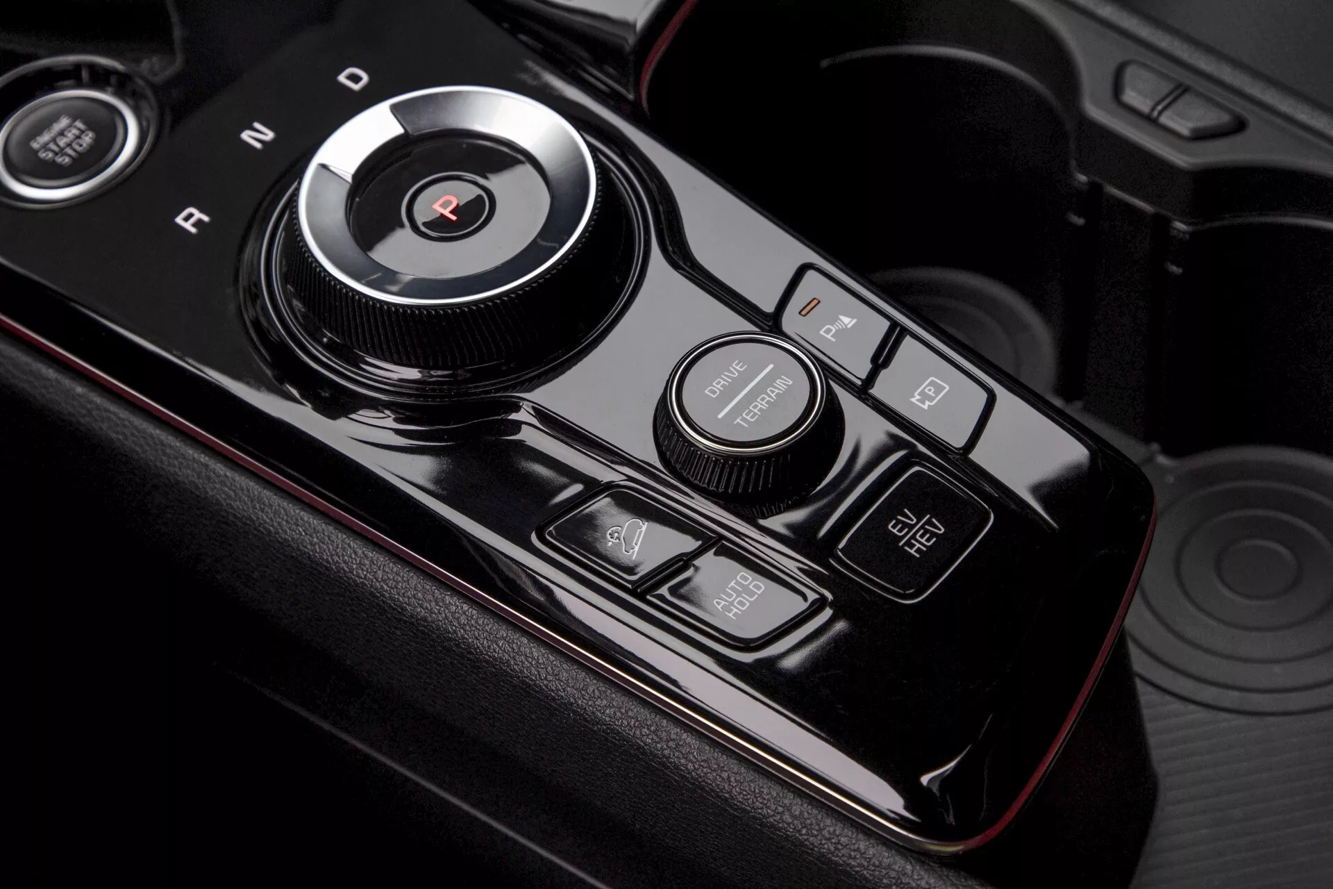 Detailní záběr na středovou konzoli Kia Sportage (NQ5) 2022 ukazující otočný volič rychlostí pro režimy 'P', 'R', 'N', 'D', 'S', vedlejší ovladač jízdních režimů a multifunkční tlačítka s LED indikací na lesklém černém povrchu s červenou dekorací