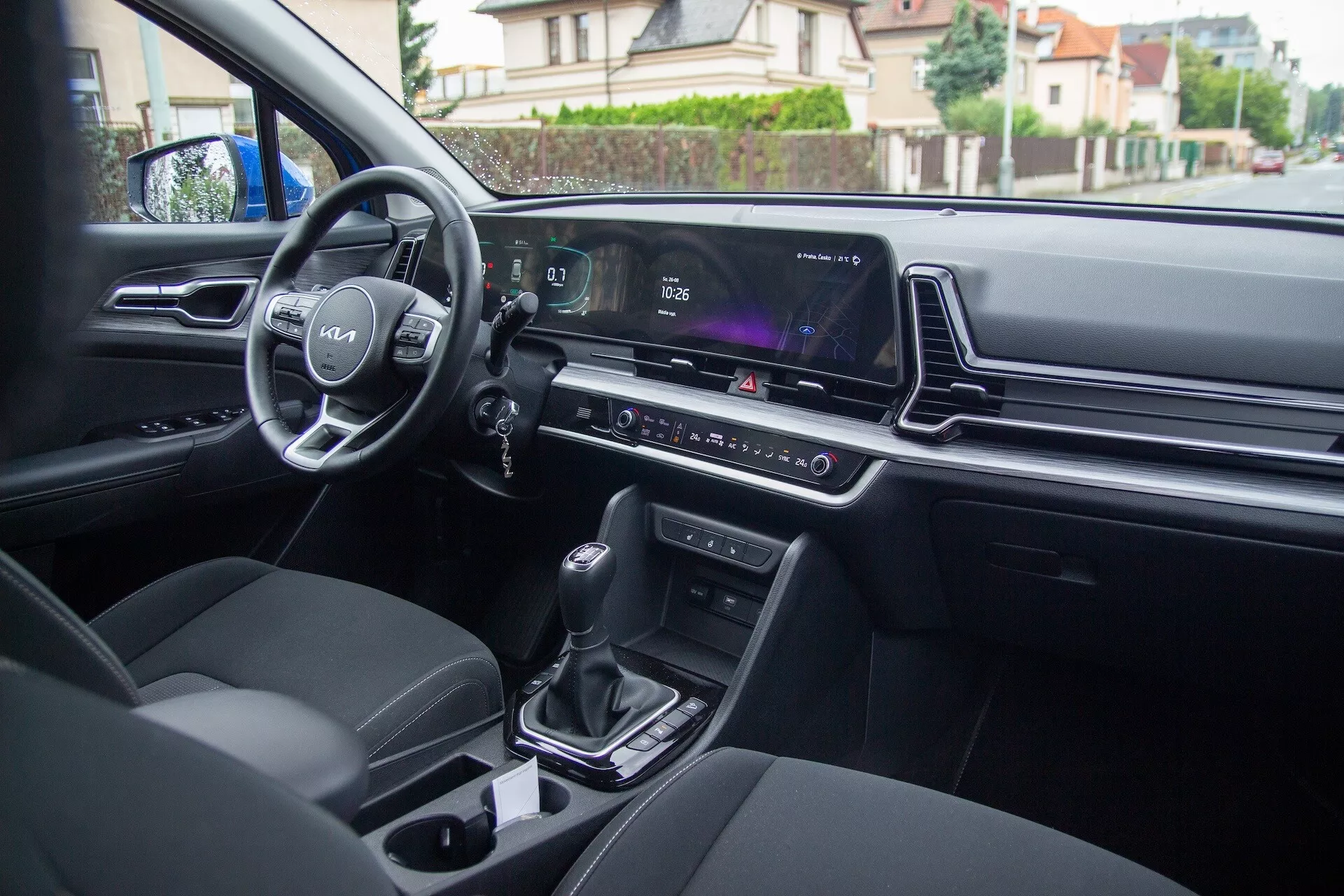 Interiér Kia Sportage 2022 z pohledu ze zadního sedadla ukazující ovládací prvky na dveřích řidiče, volant s logem a namontovanými ovladači, digitální přístrojový panel, obrazovku infotainmentu, klimatizaci, automatickou řadicí páku, držáky na nápoje a moderně čalouněná sedadla