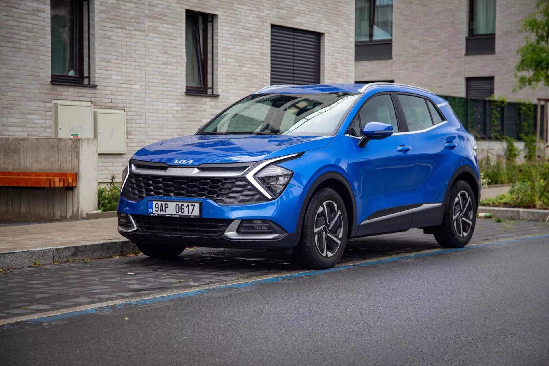 Modré kompaktní SUV crossover Kia Sportage 2022 zaparkované na mokré ulici, prezentující svůj dynamický přední úhel s LED denními světly, sochařsky modelovanou kapotou a slitinovými koly.