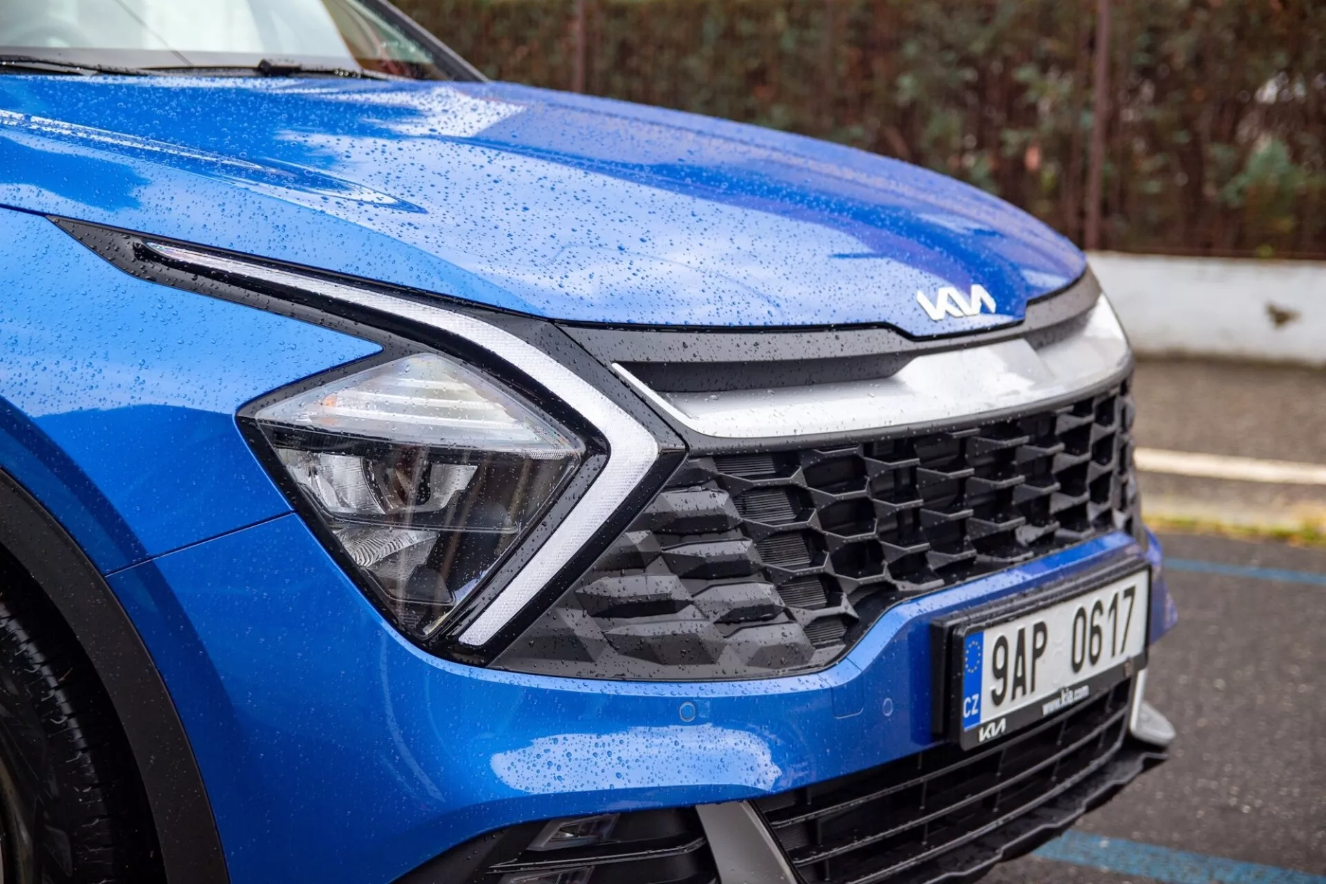 Detailní pohled na modrý Kia Sportage 2022 zaměřený na přední levý čtvrtletí za mokrých podmínek, zdůrazňující dynamický design kapoty, LED světlomety, mřížku ve tvaru včelího plástve a pokročilou senzorovou technologii