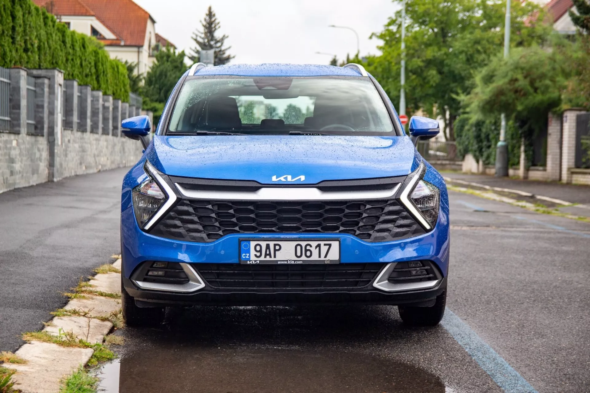 Pohled z předního úhlu na modrý Kia Sportage 2022 zaparkovaný na předměstské silnici, předvádějící jeho moderní mřížku, elegantní světlomety, chromované logo Kia a dynamický design přední části