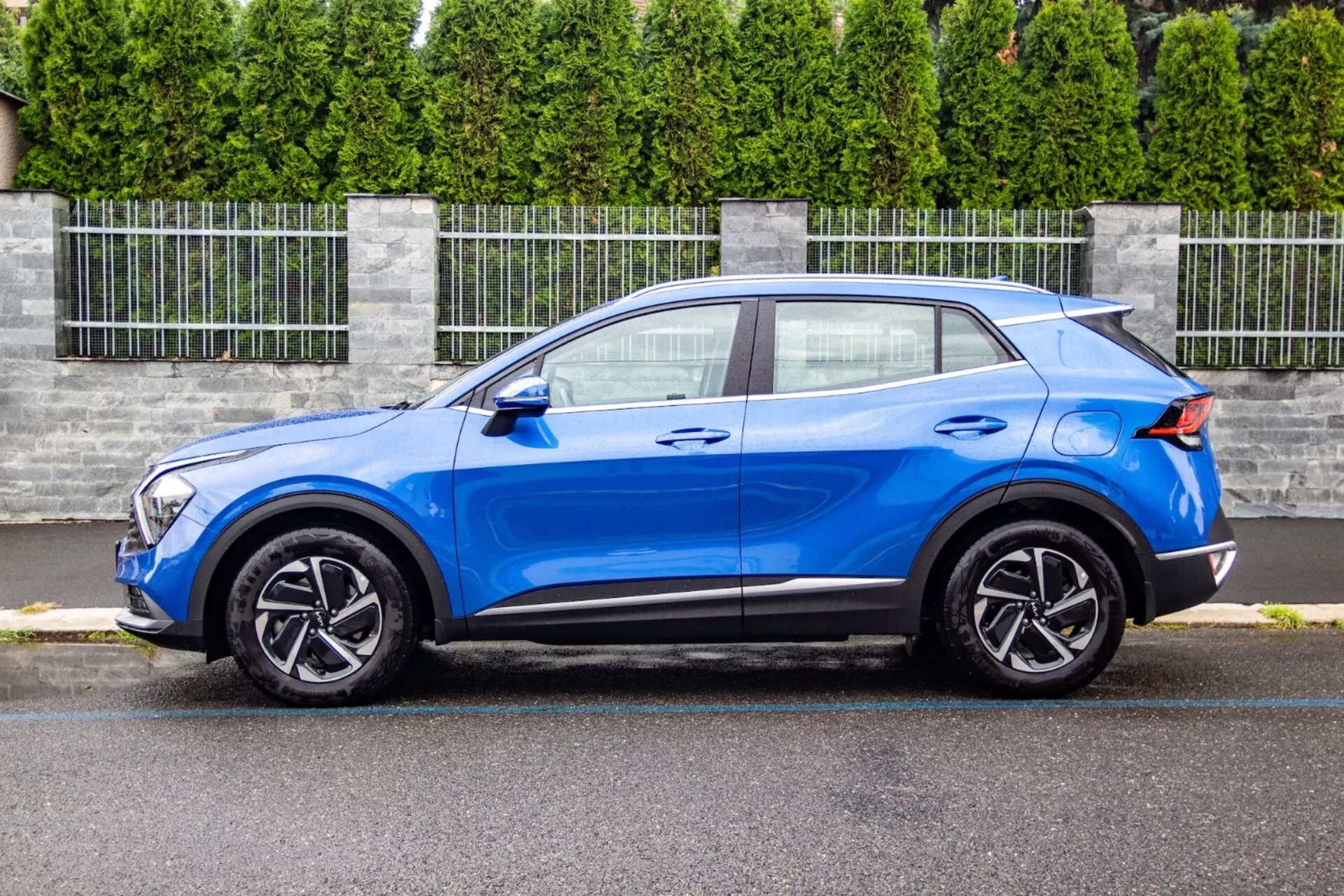 Crossover SUV Kia Sportage 2022 v jasně modré barvě předvádí svůj štíhlý, kompaktní design s kupé podobným sklonem střechy, profilovanou kapotou, jednotkami LED světlometů, pěticípými slitinovými koly a aerodynamickými vylepšeními, jako je zadní spoiler, v předměstském prostředí.