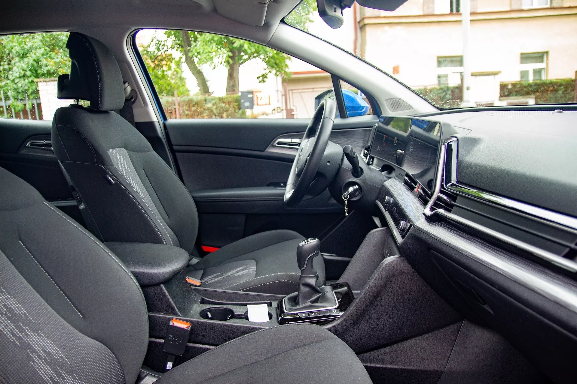 Interiér Kia Sportage 2022 z pohledu zadního pasažéra ukazující přední sedadlo spolujezdce, centrální konzoli s automatickou řadicí pákou, volant na levé straně a velkou informační obrazovku na palubní desce