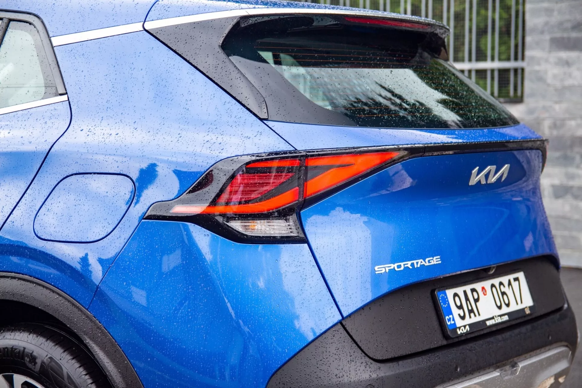 Částečný pohled na modrý Kia Sportage 2022 zdůrazňující sofistikované Z-tvarované LED zadní světlo a detaily zadní části, včetně odznaku modelu, SPZ a barvy poseté kapkami deště