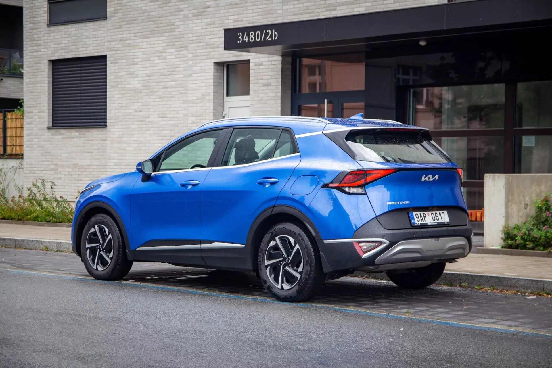 Moderní crossover SUV Kia Sportage 2022 zaparkovaný na městské ulici, zobrazující zadní tříčtvrteční pohled s výraznými zadními světly, sportovním spoilerem na střeše, slitinovými koly s více paprsky, živou modrou barvou a potenciálně moderními technologickými funkcemi.
