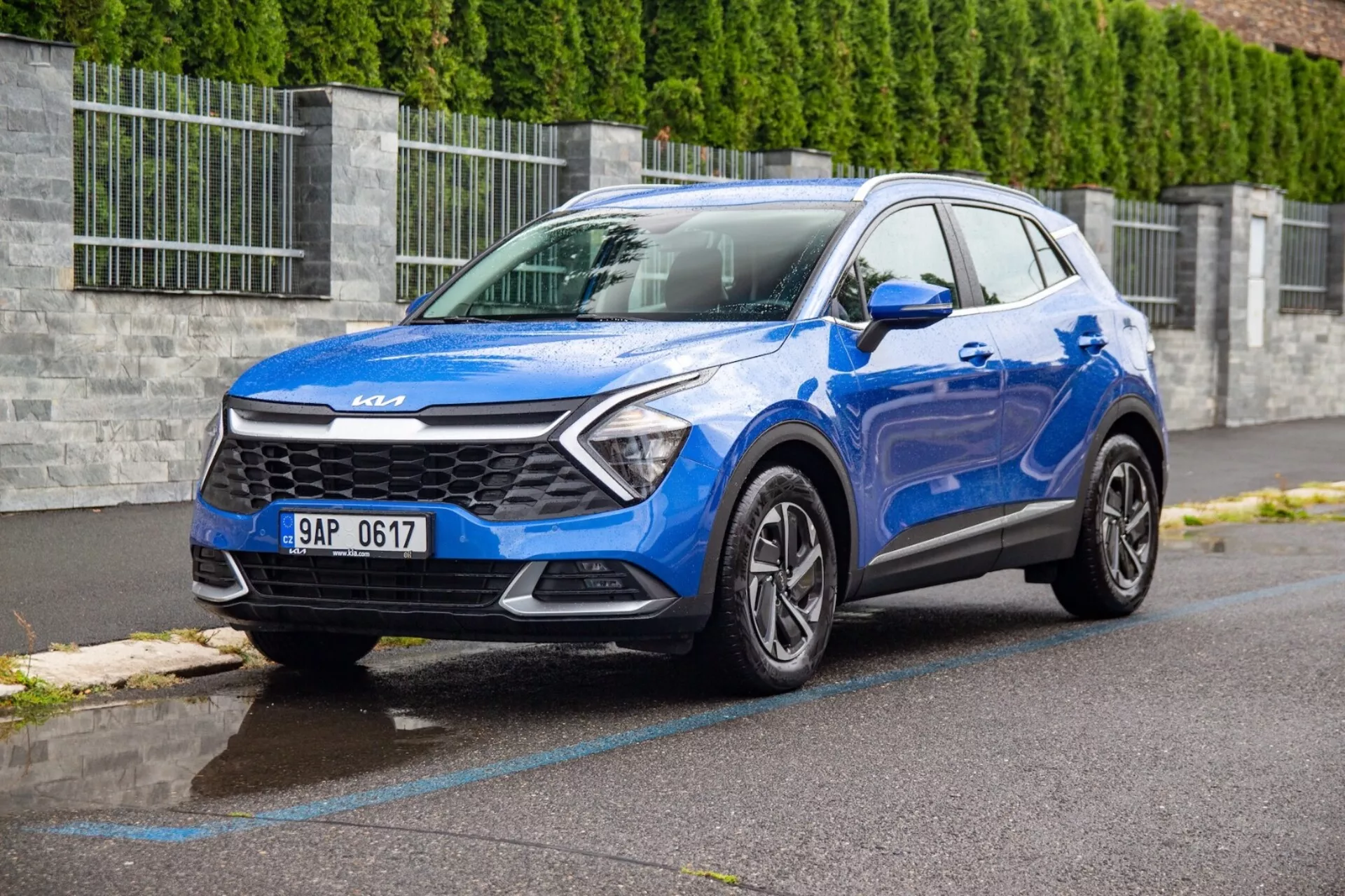 Modrý kompaktní SUV Kia Sportage 2022 zaparkovaný na mokré asfaltové ulici zobrazuje přední mřížku, světlomety, nárazník, kapotu a profil řidičovy strany z úhlu tři čtvrtiny zepředu s nízkým pohledem, který zdůrazňuje design vozidla a technologické funkce