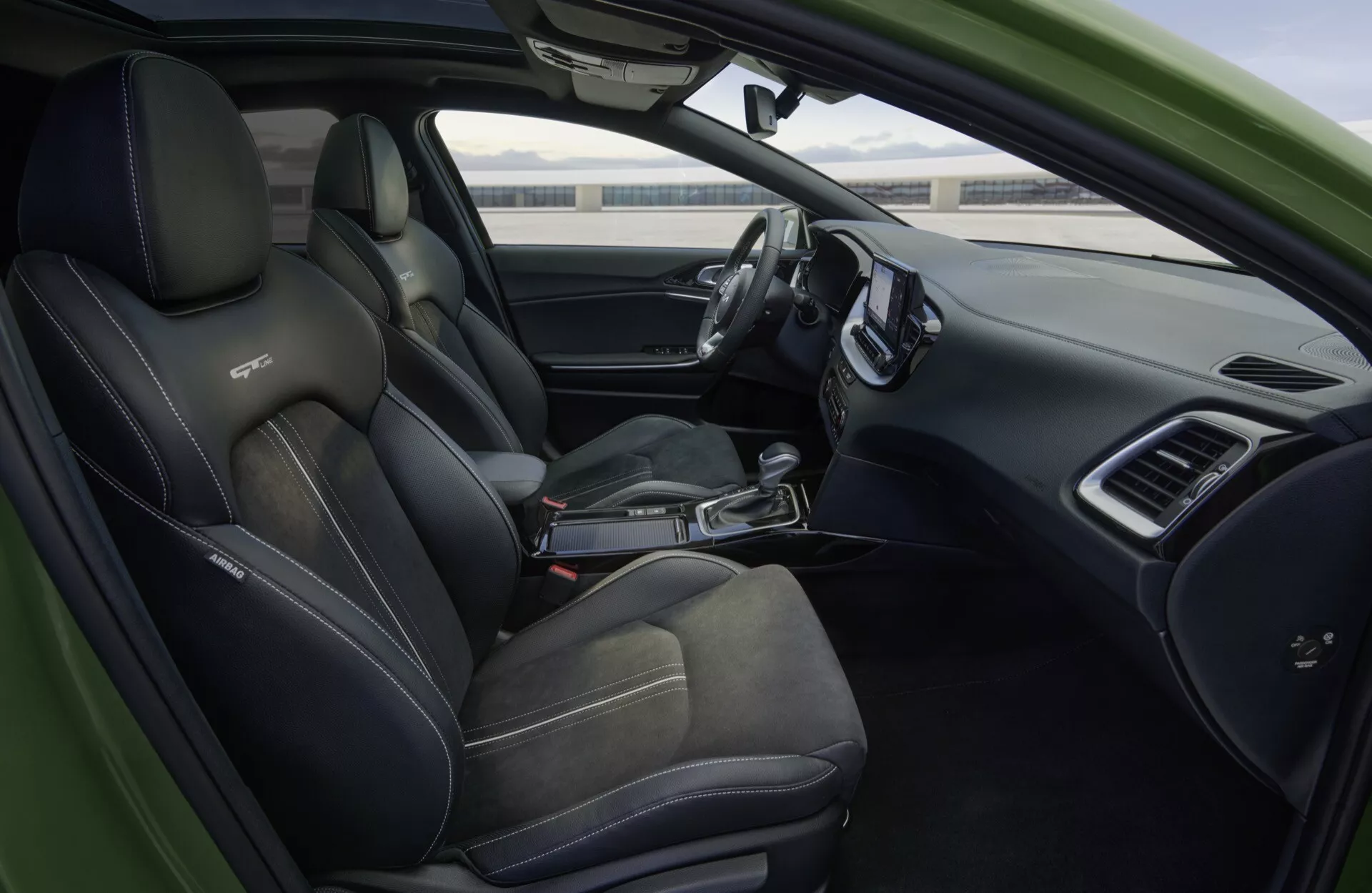 Interiérový pohled na Kia XCeed 2022 CD-facelift s sportovními černými sedadly s kontrastním prošíváním, volantem s plochým dnem obaleným kůží s multifunkčními ovládáními, elegantní palubní deskou s velkou obrazovkou infotainmentu, digitálním přístrojovým panelem, voličem automatické převodovky a ovládáními na dveřích řidiče, což vše ztělesňuje moderní automobilovou technologii a luxusní funkce.