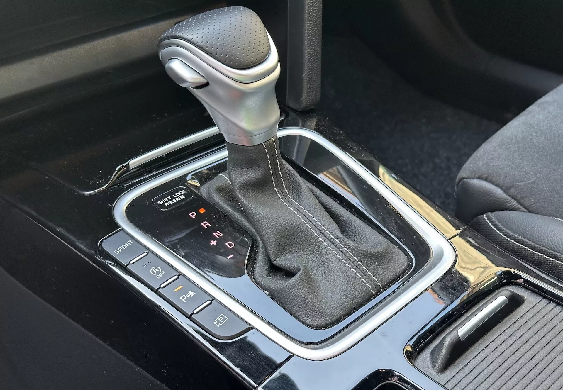 Detailní pohled na automatickou řadicí páku Kia XCeed 2022 s obalem z pravé kůže a chromovaným lemem, umístěnou na vysoce technologické středové konzoli