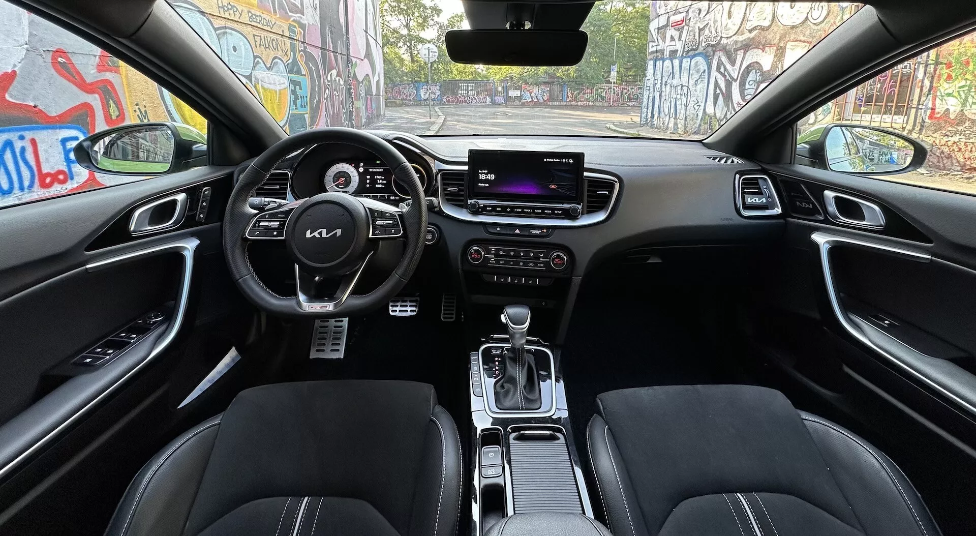 Interiér Kia XCeed 2022 z pohledu zadních sedadel, ukazuje volant s ovládacími tlačítky, digitální přístrojový panel, centrální informační obrazovku, manuální ovládání klimatizace, přední sedadla s koženým čalouněním, volič převodovky v poloze park, a pokročilé funkce jako ovládání jízdních režimů a zrcátko se zatmíváním, vše v tmavém barevném schématu.