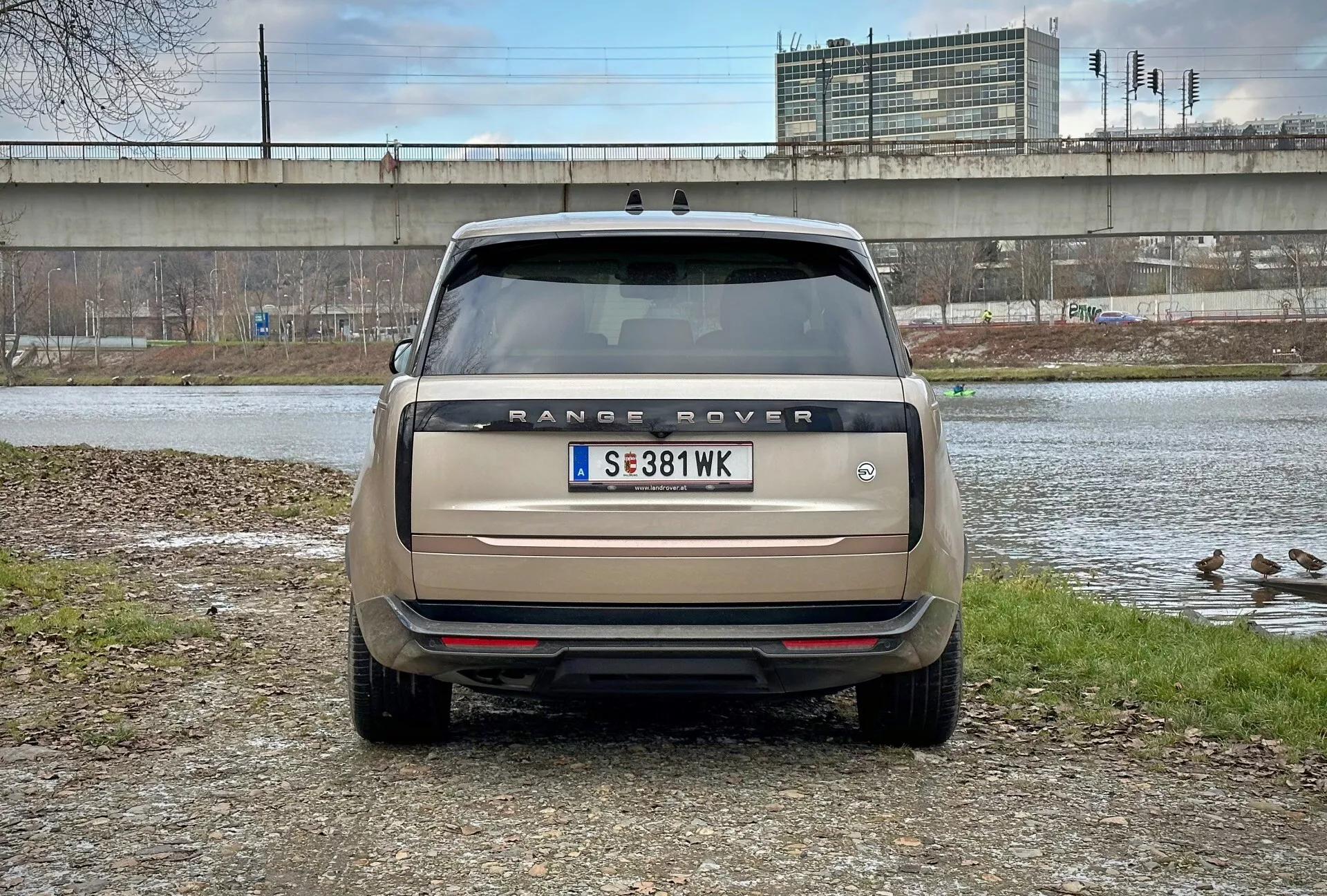 Zadní pohled na Range Rover 2021 s přímým úhlem pohledu ve výši očí, parkovaný u vodní plochy s kachnami a nadjezdem v pozadí, zobrazující název modelu, zadní světla, parkovací senzory a dvojitý výfukový systém v matné béžové barvě
