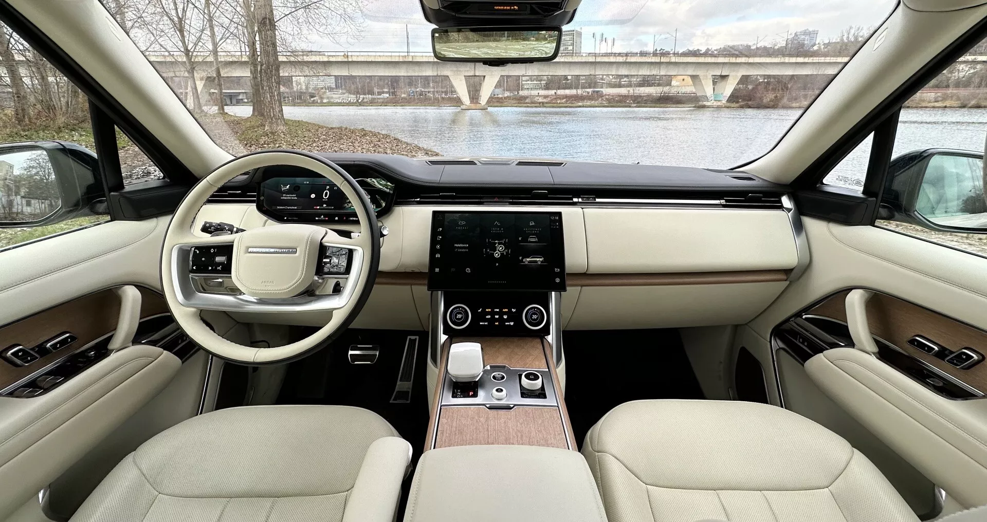 Interiér Land Rover Range Rover 2021 z pohledu ze zadních sedadel směrem k palubní desce s symetrickým designem, volantem s multifunkčními tlačítky, dotykovým systémem infotainment, digitálním ovládáním klimatizace, luxusním koženým čalouněním s dřevěnými akcenty a pokročilými technologickými funkcemi