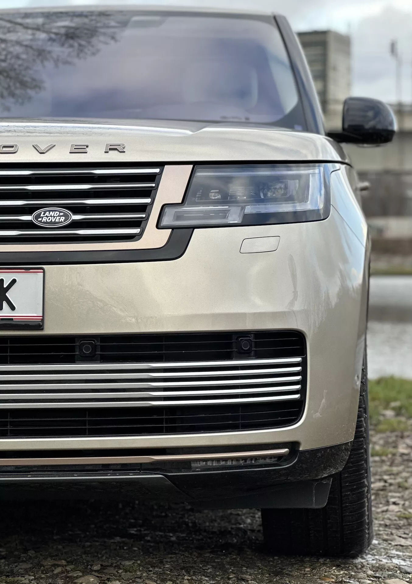 Detailní záběr zlatého Land Roveru Range Roveru 2021 na přední levou část vozidla, zdůrazňující výšku a postoj vozidla s důrazem na kapotu, mřížku, LED světlomet, nárazník a celoroční pneumatiku, naznačující luxus a schopnost jízdy v terénu.