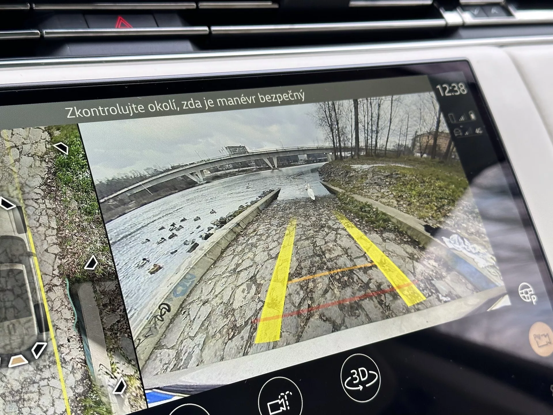 Displej informačního systému Land Rover Range Rover 2021 ukazující zadní pohled z kamery pro couvání s dynamickými vodicími liniemi na parkovišti u vodní plochy, detaily s vegetací, ikonami asistence při parkování a bezpečnostním upozorněním