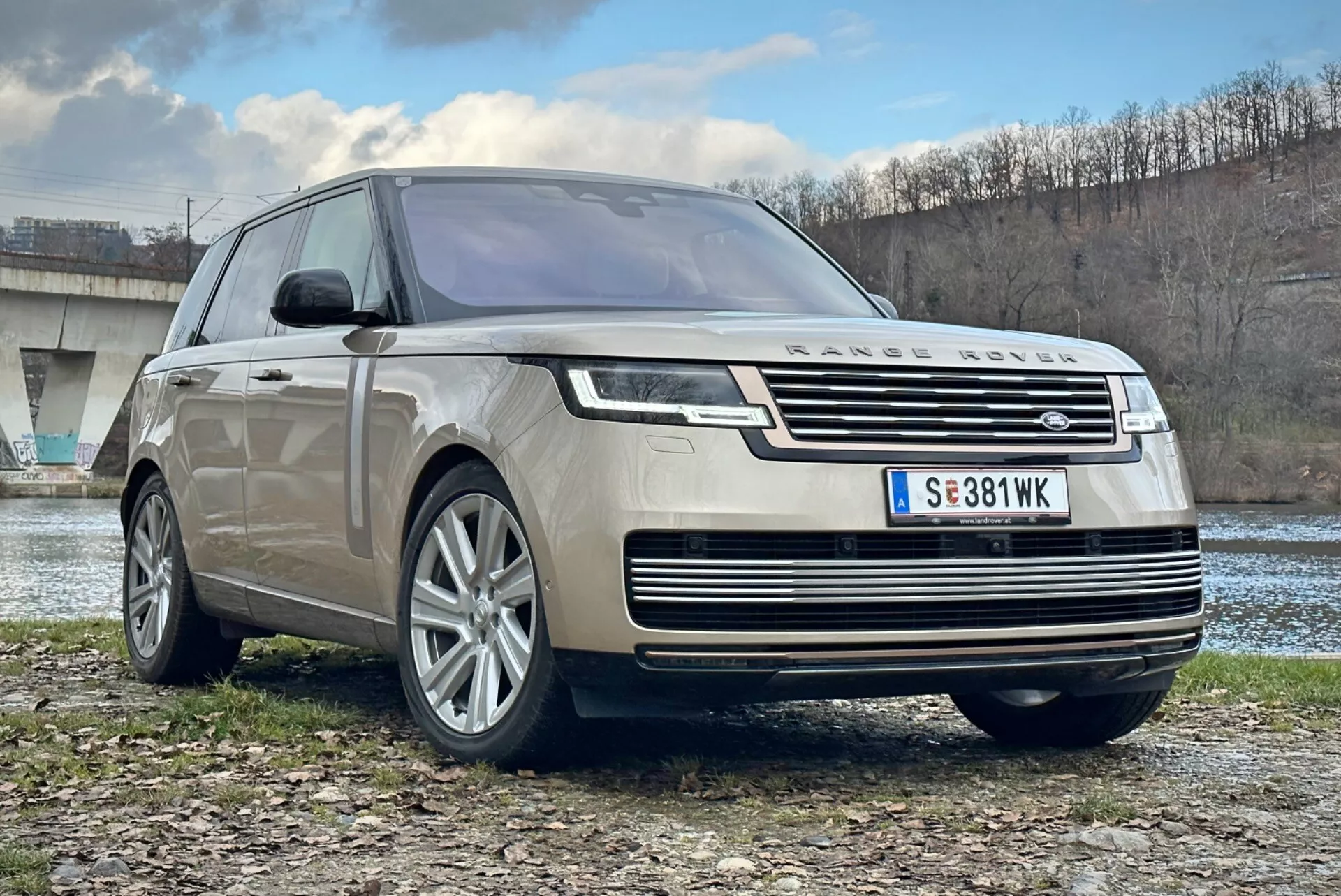 Luxusní SUV Land Rover Range Rover 2021, pohled zepředu a tříčtvrteční úhel ukazující chromovou mřížku, LED světlomety, slitinová kola s více paprsky a aerodynamický design, zaparkované v exteriéru poblíž řeky a mostu