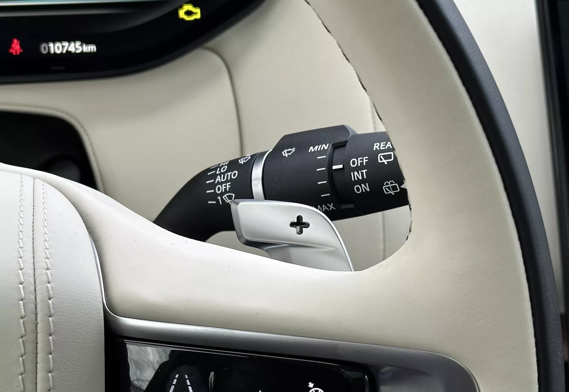 Detailní pohled do interiéru Land-Rover Range-Rover 2021 na sloupek řízení, páčku ovládání stěračů čelního skla s více nastaveními včetně OFF, INT, ON, regulátory rychlosti a AUTO pro automatické čidlo deště, plus možnost zadního stěrače. Viditelná část přístrojové desky zobrazuje 10745 km, vše proti pozadí prémiového černého a krémového interiéru.
