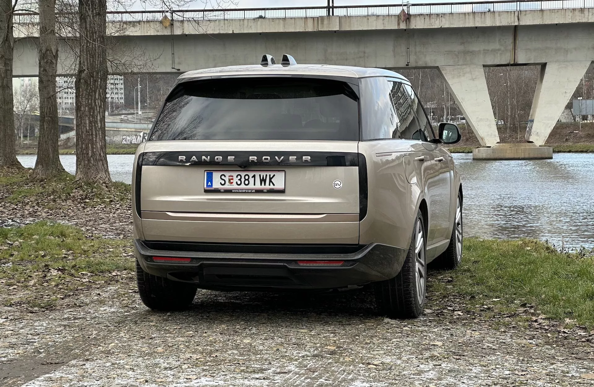 Zadní pohled na Range Rover 2021 zaparkovaný venku s vodní plochou a mostem na pozadí, ukazující LED zadní světla, dvojitou výfukovou soustavu, skla pro zvýšené soukromí a kola z lehkých slitin s více paprsky.