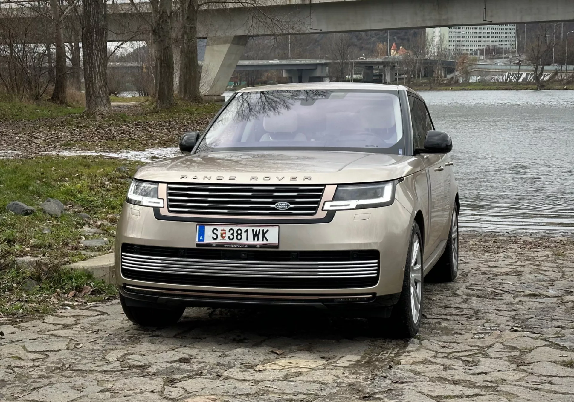 Luxusní SUV Range Rover 2021 ve stříbrné barvě, představuje výraznou přední mřížku s logem, elegantní LED denní světla, přední nárazník s parkovacími senzory a design kapoty typu clamshell na dlažební ploše u řeky