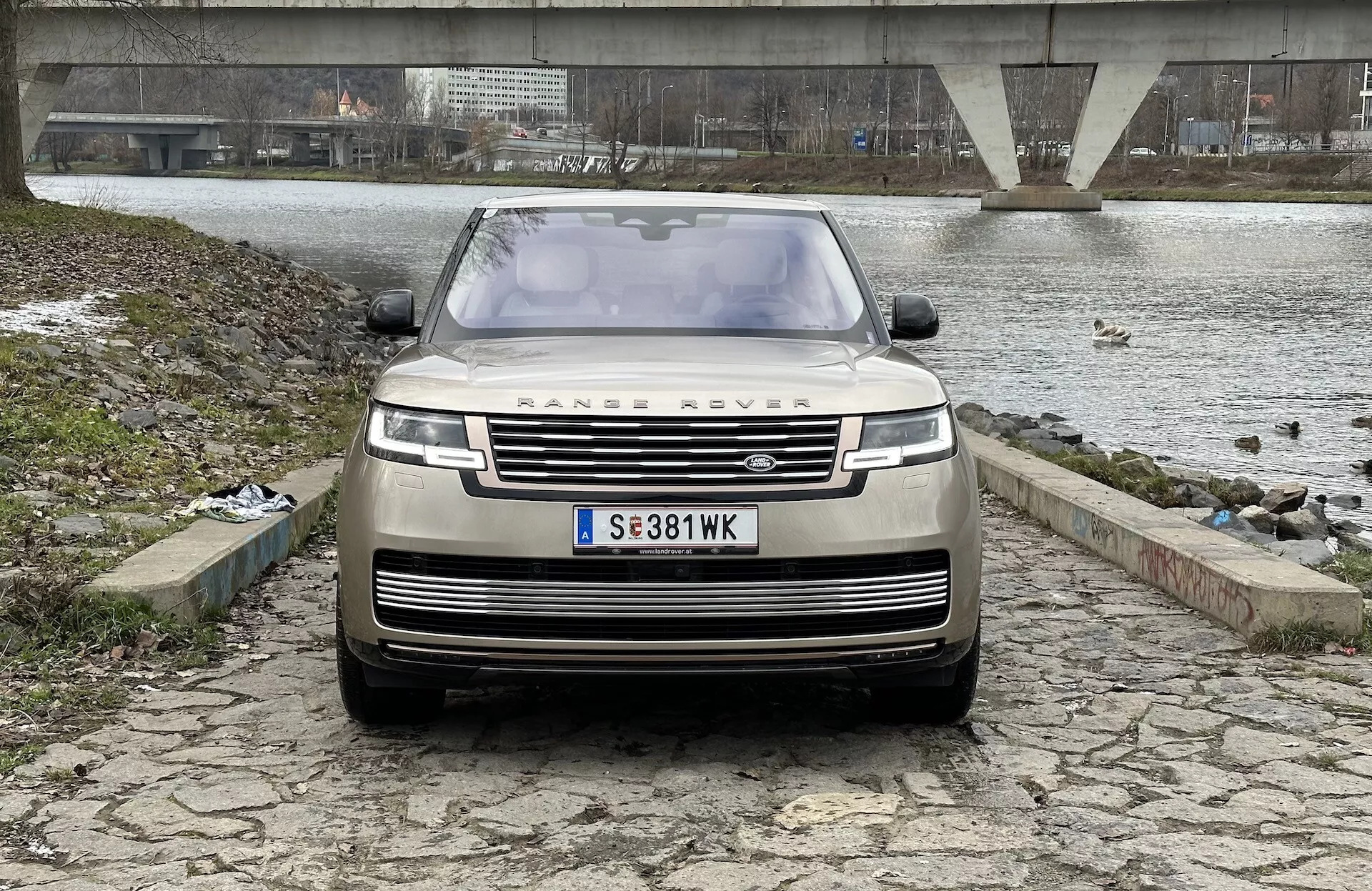 Land Rover Range Rover 2021 zobrazený z předního, mírně nízkého úhlu zdůrazňujícího mřížku, přední nárazník a kapotu, s ikonickým emblémem RANGE ROVER, moderními LED světlomety a pokročilou technologií asistence řidiče, na pozadí řeky nebo jezerní krajině, mostu a zamračené oblohy