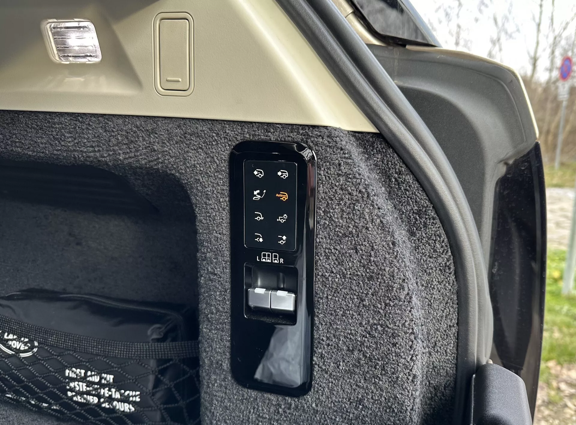 Detailní pohled na elektronický ovládací panel kufru ve vnitřní části Land Rover Range Rover 2021, zobrazující tlačítka pro operace jako elektronické otevírání a zavírání, ovládání nákladového krytu, potenciálně světlo kufru nebo zásuvku, nastavení zadních sedadel z kufru a nouzové uvolnění, umístěné proti kobercové boční stěně kufru a světlému stropu s osvětlovacím tělesem nákladu.