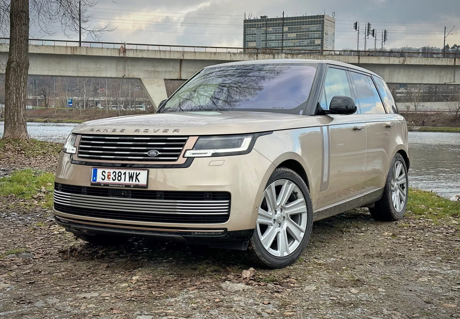 Luxusní SUV Land Rover Range Rover 2021 zaparkované na smíšeném terénu země a trávy s betonovou konstrukcí a vegetací v pozadí, ukazující pohled z předního úhlu zdůrazňující LED světlomety, slitinová kola a moderní aerodynamické prvky