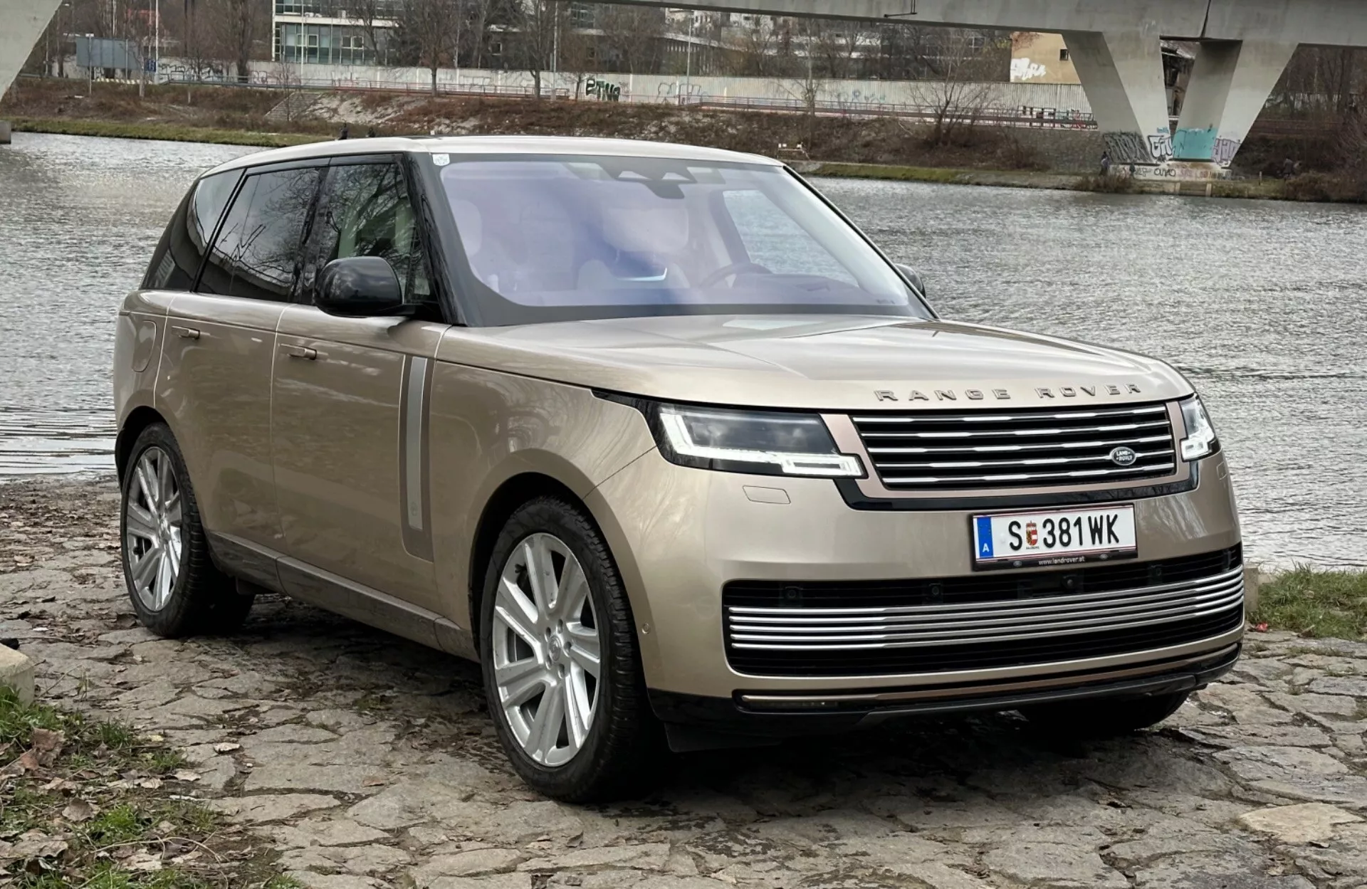 Luxusní SUV Land Rover Range Rover ročník 2021 zaparkované na břehu řeky s mostem na pozadí, předvádějící jeho moderní design a technologické prvky včetně LED světlometů, přední mřížky s emblémem, vícepaprskových slitinových kol a panoramatické střešní okno.