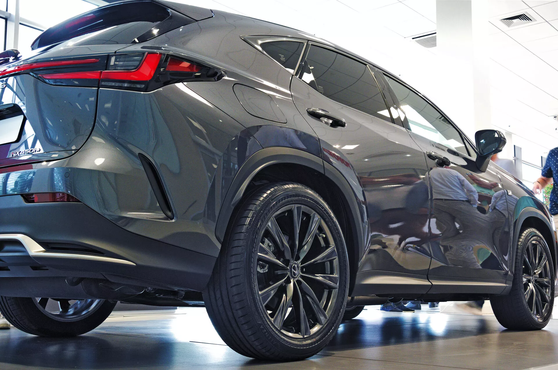 Zadní tříčtvrtinový úhel vozu Lexus NX 2021 SUV ukazující boční profil a zadní část s velkými vícepaprskovými litými koly, nízkoprofilovými pneumatikami, ostrými zadními LED světly, metalickým lakem, tónovanými okny, integrovaným zadním spoilerem a naleštěným showroomovým prostředím zdůrazňujícím moderní estetiku a luxusní prvky