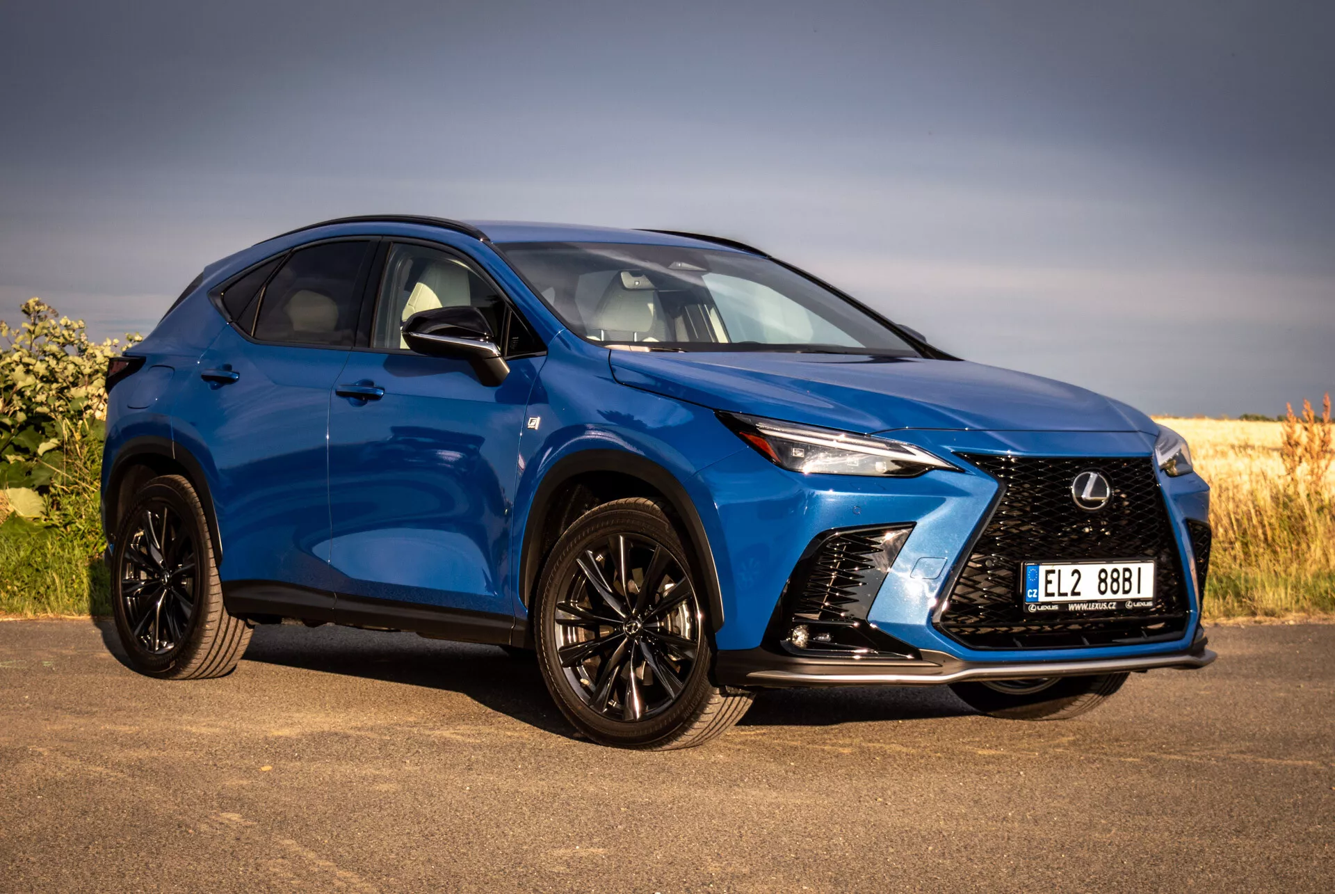 Modré SUV Lexus NX 2021 ve čtvrtém předním pohledu zdůrazňující jeho agresivní mřížku, ostrá LED světlomety, černé slitinové kola s nízkoprofilovými pneumatikami a lakovaná boční zrcátka na pozadí přírody