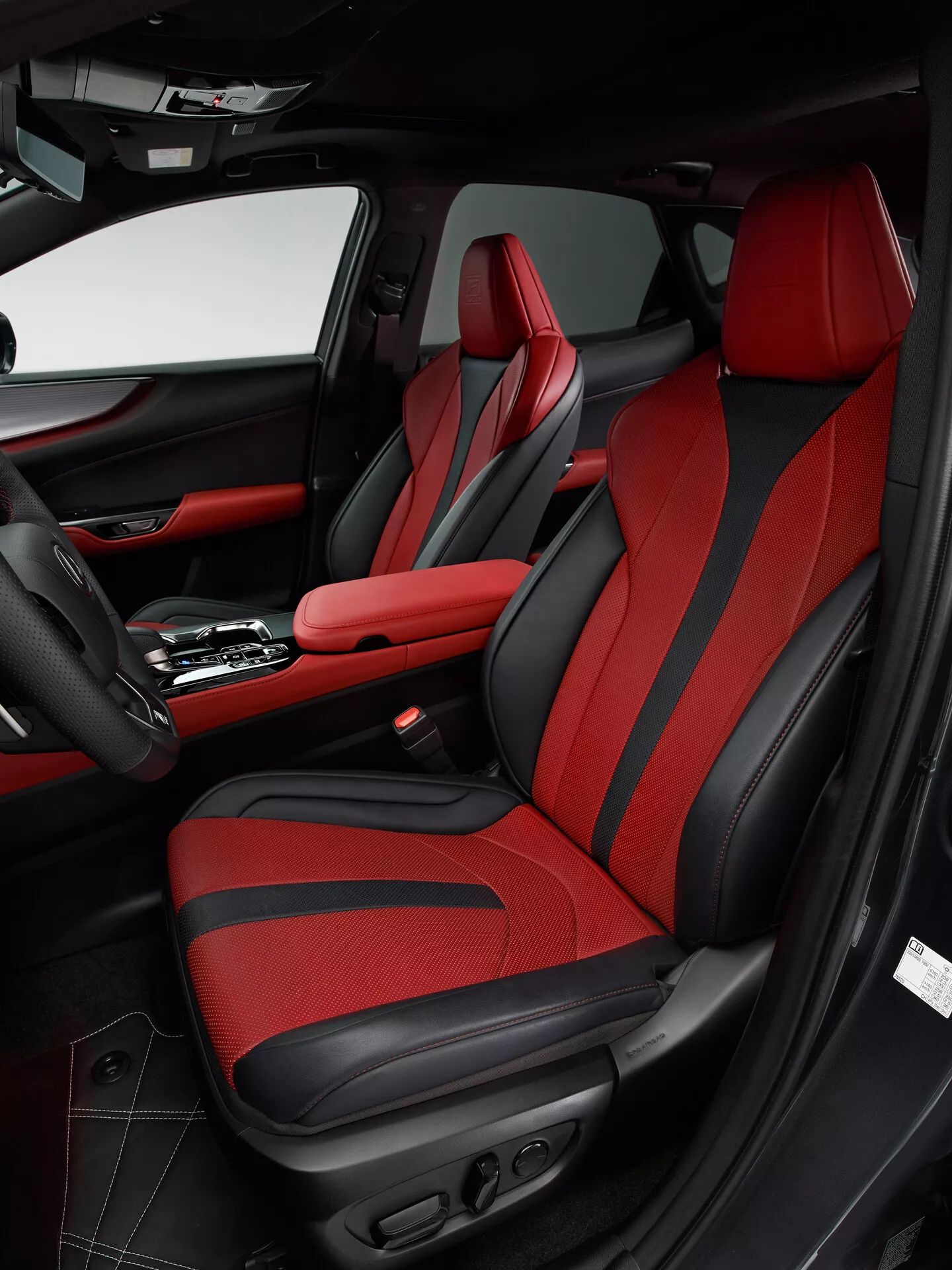 Záběr interiéru Lexus NX 2021, zobrazující oblast předního spolujezdce z úhlu od strany řidiče. Obrázek zdůrazňuje sedadlo spolujezdce s výrazným červeným čalouněním a černými akcenty, sportovním designem s vylepšeným bočním vedením a ovládáním nastavení. Dveřní panel odpovídá černočervenému barevnému schématu, obsahuje kůži, tkaninu, kapsu na odkládání, mřížku reproduktoru a různé ovládací prvky. Středová konzola ukazuje automatickou řadící páku a centrální opěrku sředním červeným prošitím, odrážející luxusní a sportovní design vozu.