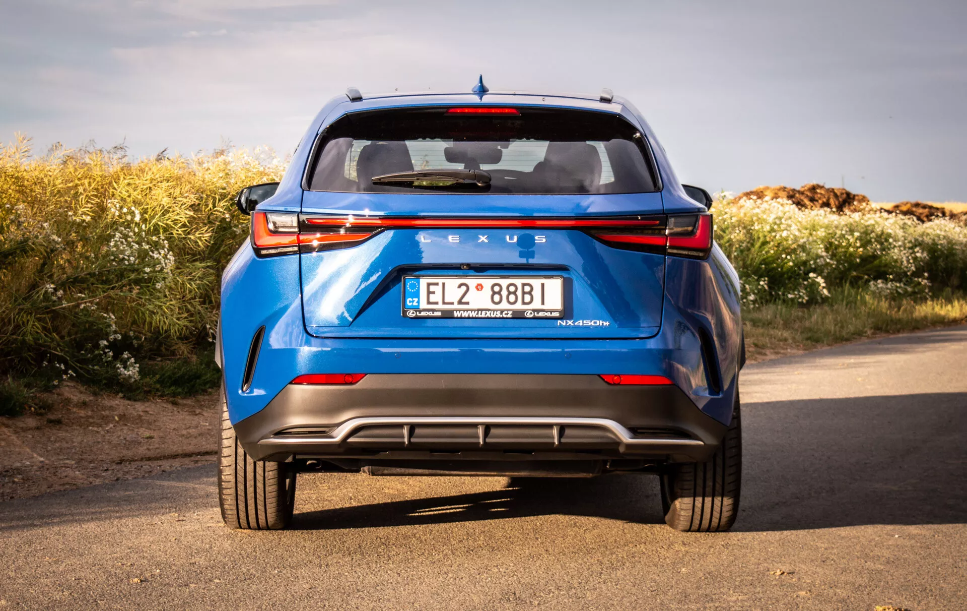 Zadní pohled na modrý Lexus NX 2021 z nízkého úhlu zdůrazňující pevnou konstrukci a designové prvky vozu, jako jsou LED zadní světla, zadní čelní sklo, spoiler, výrazné označení, zadní nárazník s difuzorem, parkovací senzory, odrazky a dvojité výfuky na pozadí přírody pod jasným nebem.