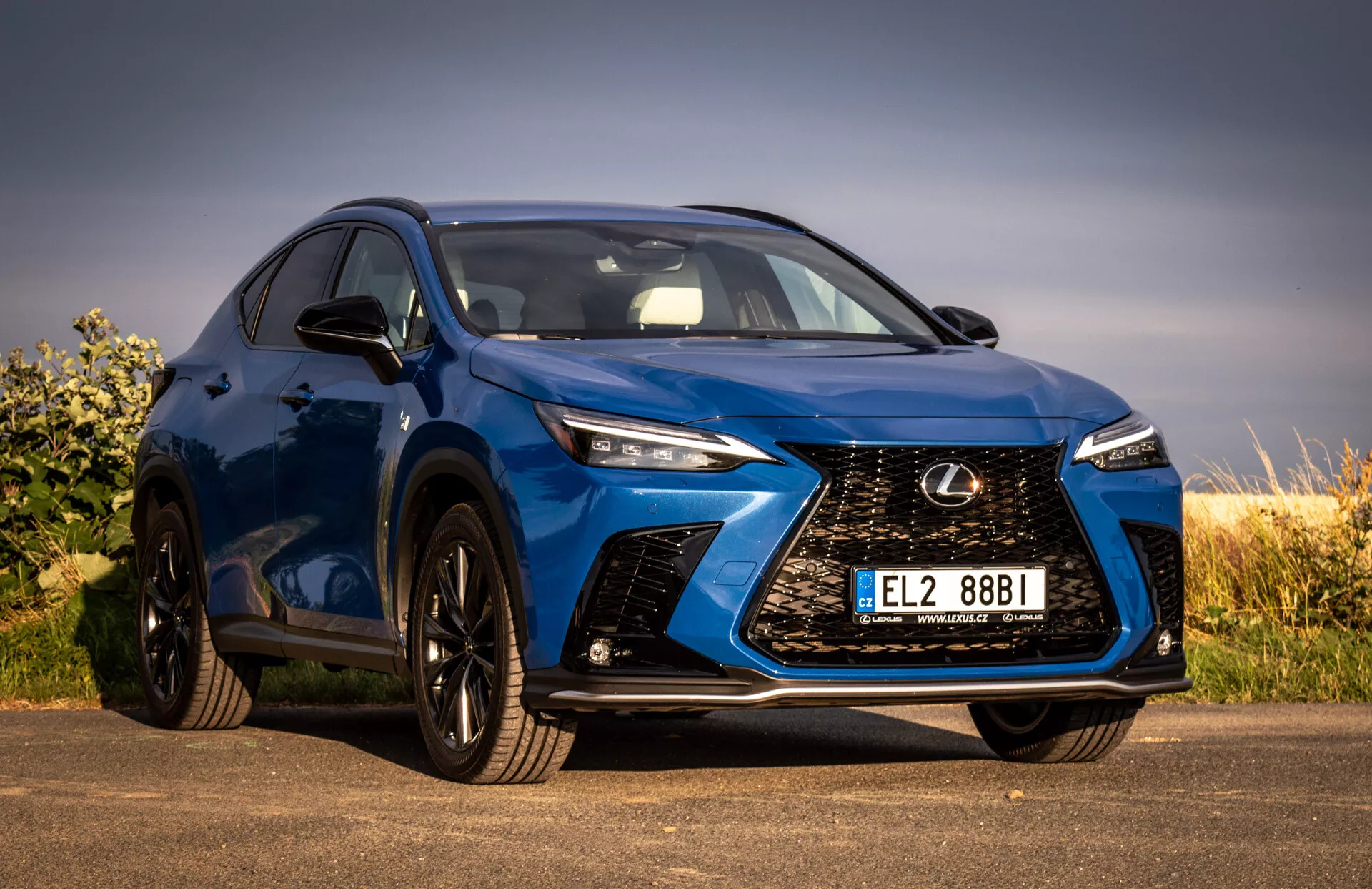Snímek z nízkého úhlu modrého SUV Lexus NX 2021 zobrazující přední mřížku se vzorem vřetene, LED světlomety, černé slitinové kola a integrované směrové signály na bočním zrcátku, zdůrazňující jeho luxusní a výkonnostní prvky.