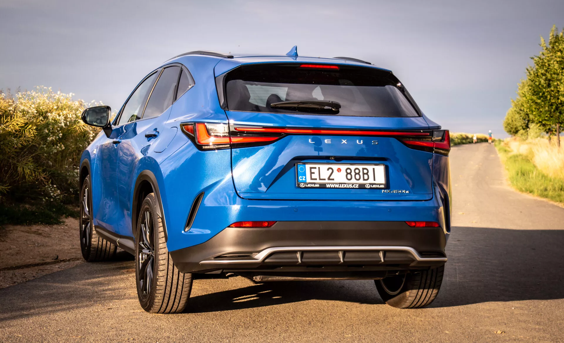 Zadní pohled na luxusní crossover SUV Lexus NX v modré barvě rok 2021, předvádějící LED zadní světla, značení Lexus, sportovní zadní nárazník s difuzorem, dvojité výfukové potrubí a další technologické a designové prvky z nízkého úhlu a trojčtvrteční perspektivy