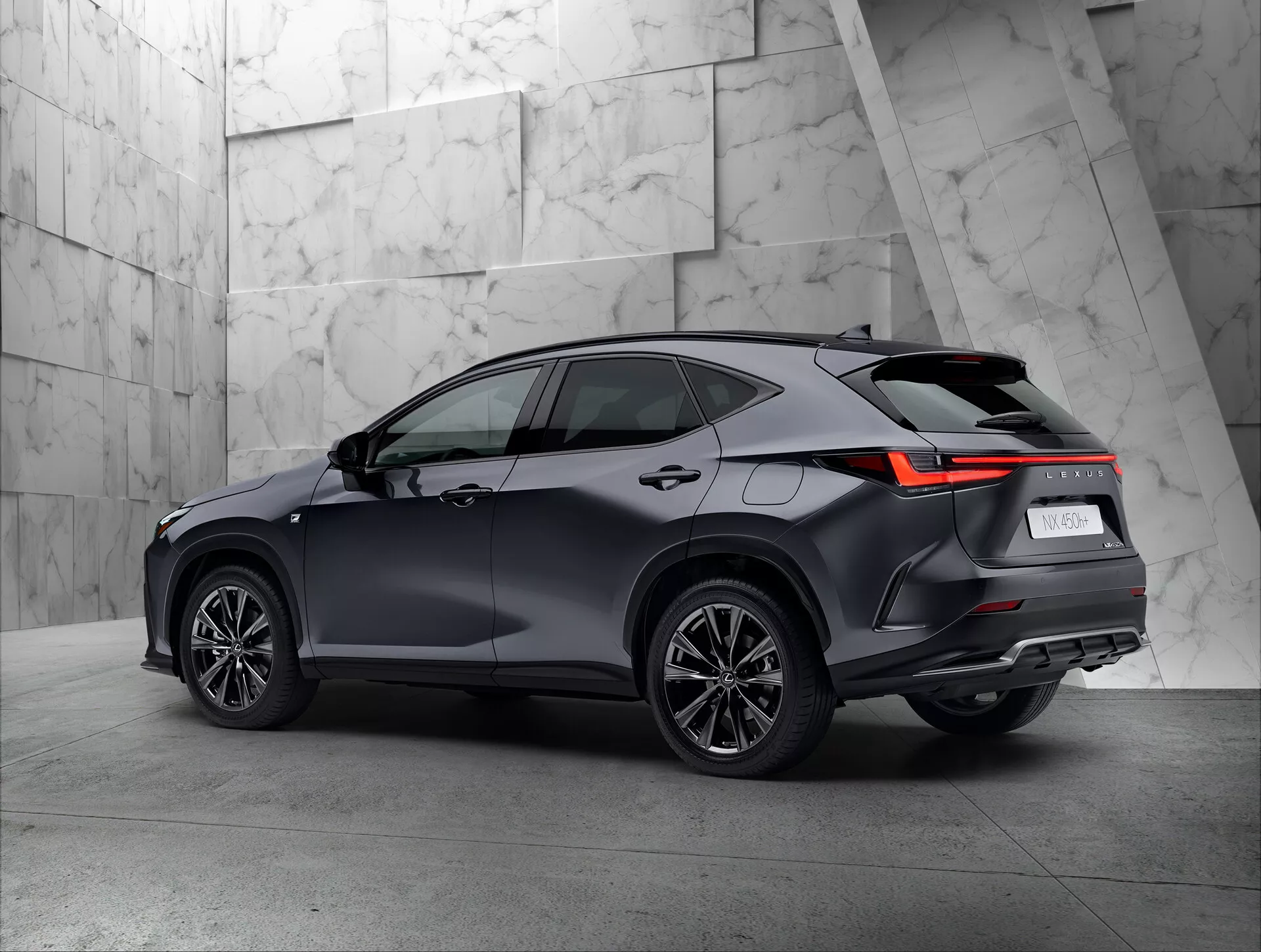 Tříčtvrteční zadní pohled na moderní SUV Lexus NX 2021 v showroomu. Vozidlo zřetelně zobrazuje pravou stranu a zadní část, včetně zadních světel s LED designem, tmavým lesklým lakem, vícepaprskovými koly a střechou s možnými střešními nosiči. Neutrální, strukturované pozadí pěkně kontrastuje se sofistikovaným designem SUV.