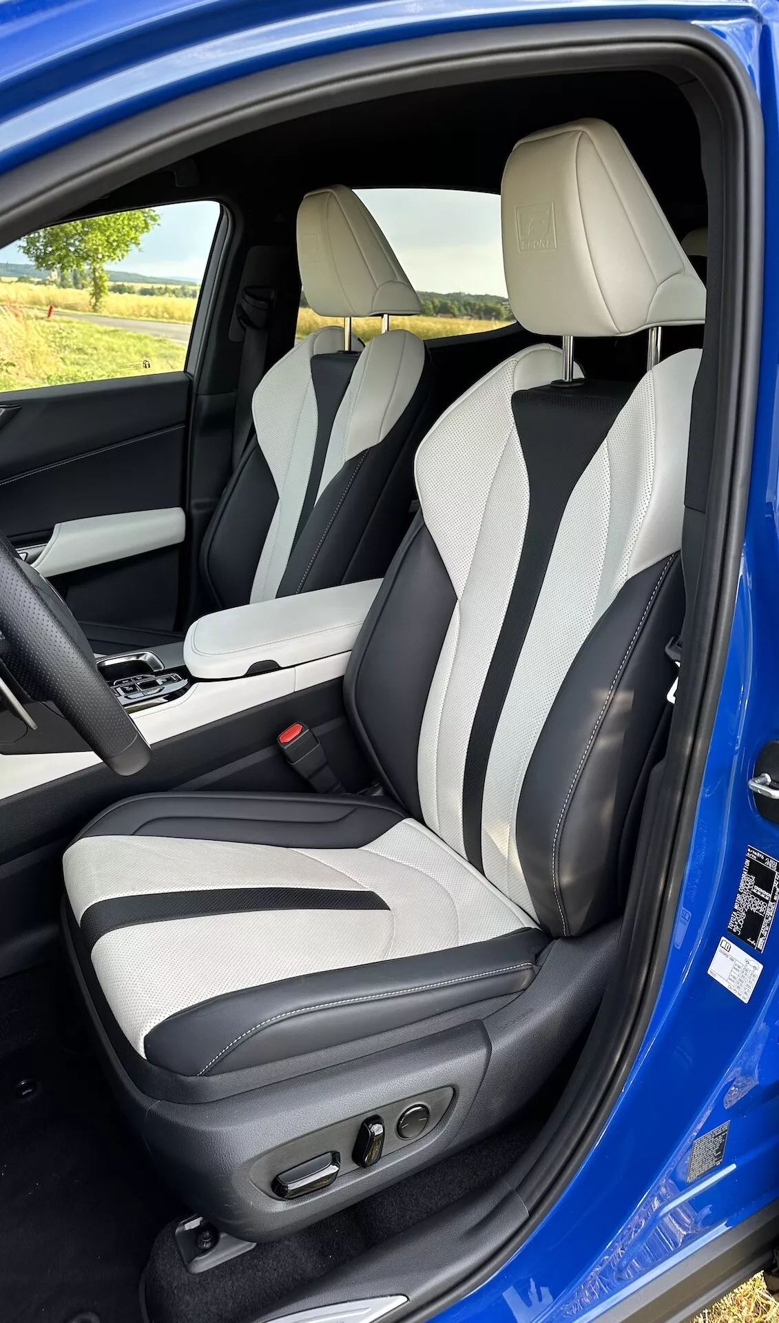 Vnitřní pohled skrz otevřené dveře řidiče na Lexus NX 2021, ukazující oblast řidiče z mírné elevace, s dvoubarevným koženým čalouněním, pokročilými ovládáními sedadla řidiče, bočním vyztužením, integrovanými opěrkami hlavy a centrální konzolou s automatickou pákou
