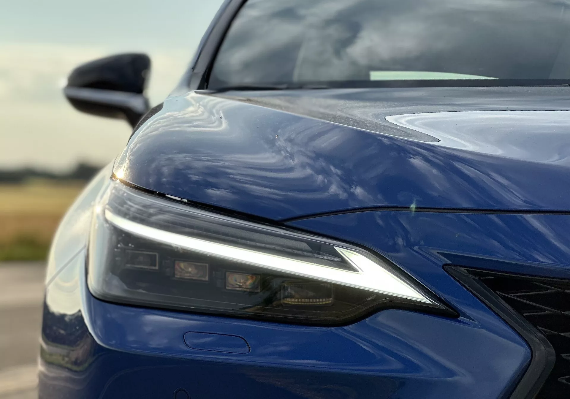 Detailní pohled na levý přední čtvrtletí Lexusu NX 2021 s LED světlometem, tvarovanou kapotou, předním nárazníkem s mřížkou a lesklým tmavě modrým povrchem, zdůrazňující pokročilou technologii osvětlení a aerodynamický design.