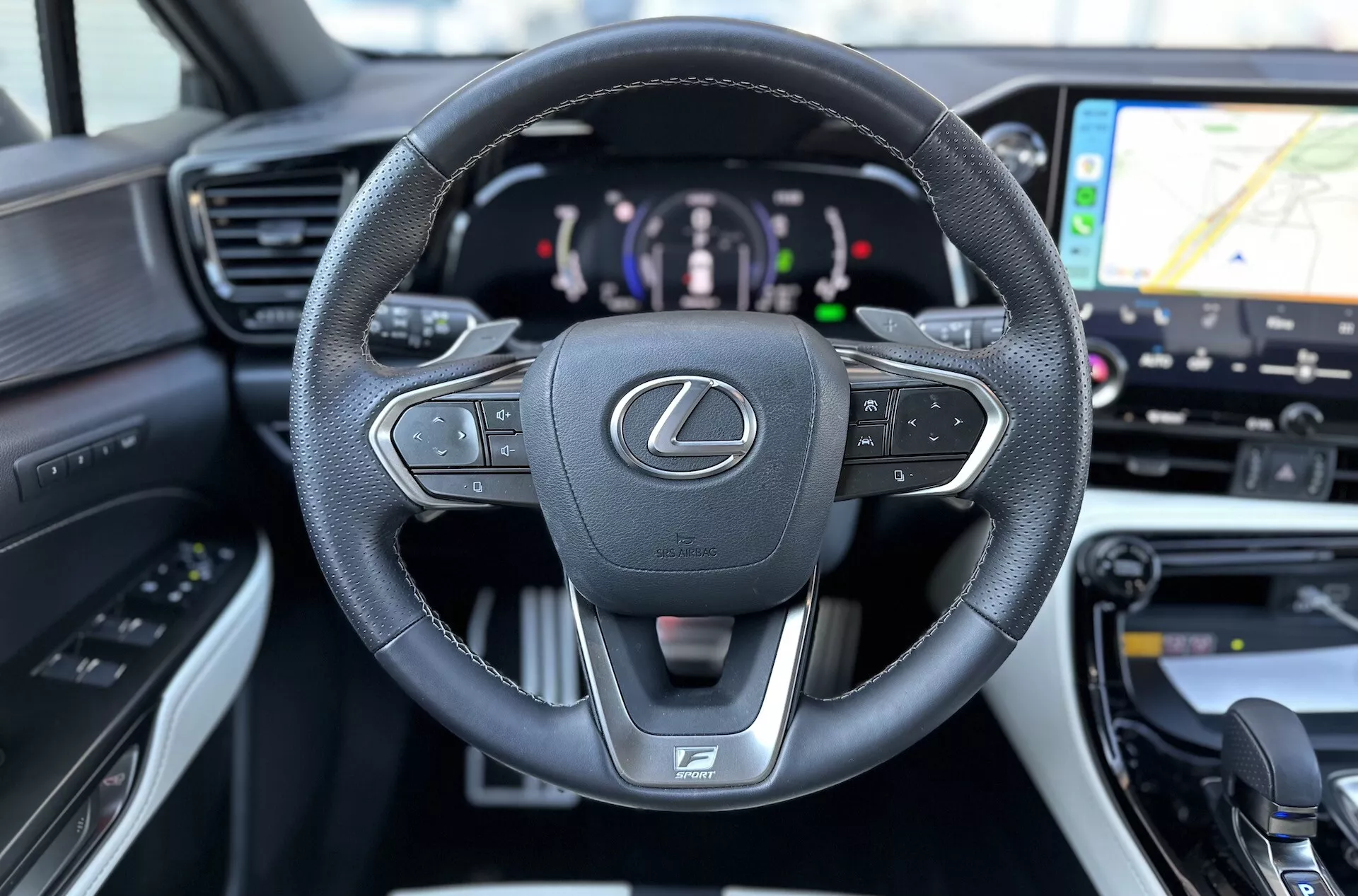Interiér Lexus NX 2021 se zaměřením na kožený, multifunkční volant s emblémem Lexus a logem F SPORT, digitální přístrojový panel naznačující hybridní pohon a náhled na GPS navigaci na systému infotainmentu