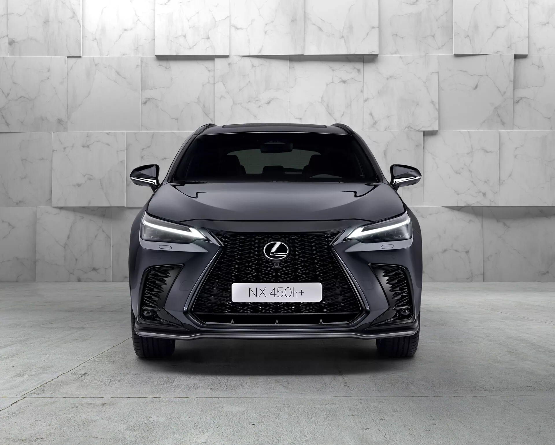 Čelní pohled na Lexus NX 450h+ z roku 2021, elegantní moderní SUV, které ukazuje svou výraznou vřetenovitou masku s mřížkovým vzorem, zúžené LED světlomety, logo Lexus s modrým obrysem a agresivní konstrukci předního nárazníku. Hybridní charakter vozu je indikovaný znakem 'h+' na masce. Studiové prostředí zvýrazňuje křivky a vlastnosti vozidla čistým, kontrolovaným osvětlením.