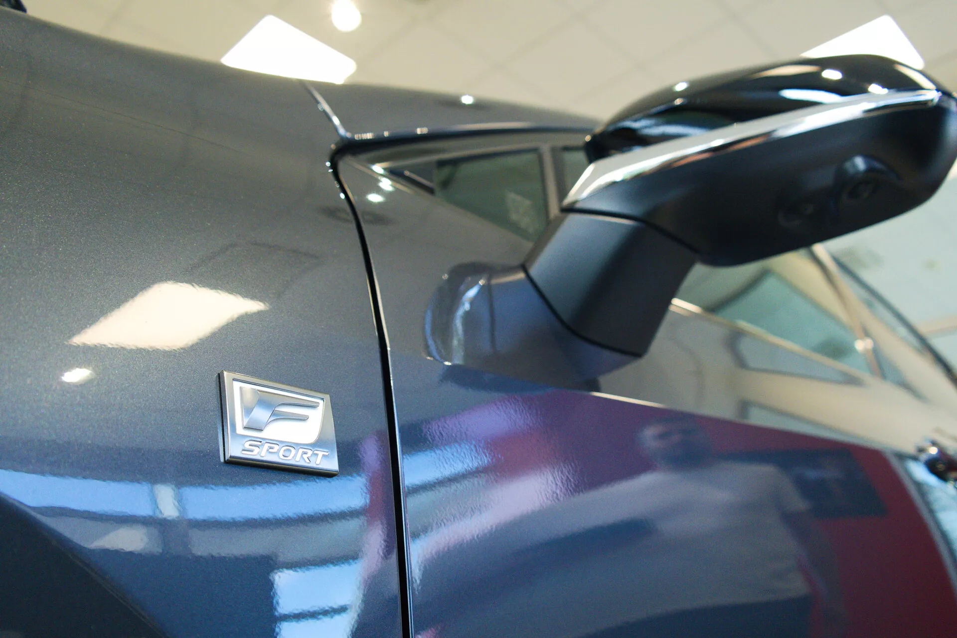 Detailní snímek vozu Lexus NX 2021, zaměřený na boční zrcátko a odznak 'Sport'. Záběr pořízený z nízkého úhlu lehce nahoru ukazuje aerodynamické zrcátko v karoserii tmavé barvy. Odznak 'Sport', indikující sportovní variantu, je umístěn nad obloukem kola s nízkou hloubkou ostrosti, což zdůrazňuje estetiku designu.