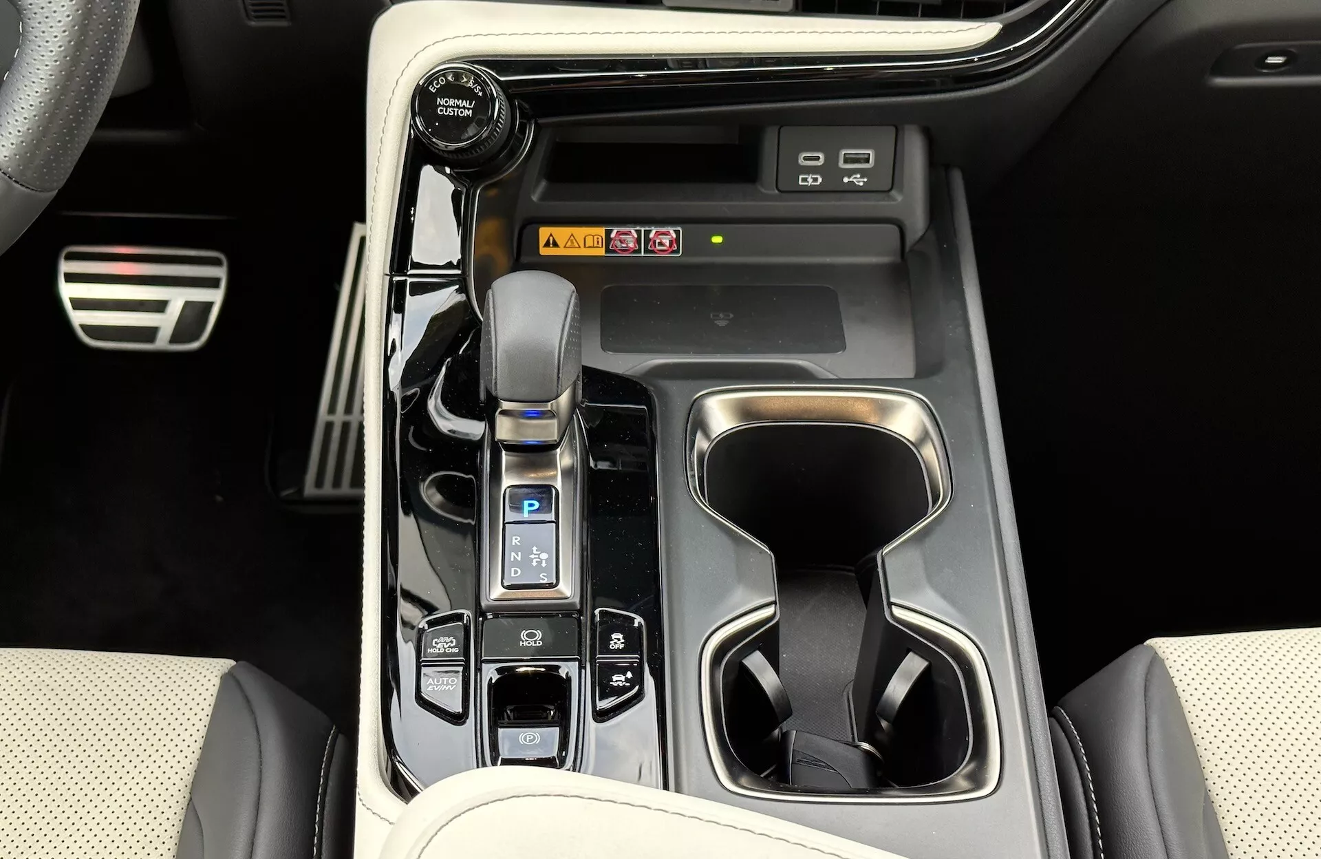 Pohled na středovou konzolu Lexus NX 2021 ukazující automatickou řadicí páku s koženým manžetou, voličem jízdních režimů, držáky na nápoje, tlačítka ovládání vozidla, potenciální zkratky pro informačně-zábavní systém a prvky luxusního interiérového designu