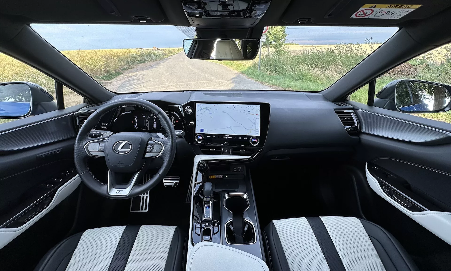 Interiér Lexus NX 2021 z pohledu řidiče s volantem uprostřed s logem Lexus a ovládacími tlačítky, velkým dotykovým displejem na palubní desce, minimalistickými ovládacími prvky klimatizace, volicí pákou automatické převodovky na středové konzole s držáky na nápoje a úložným prostorem, sedadla čalouněná dvoubarevným materiálem a částečným výhledem na venkovskou krajinu přes čelní sklo.