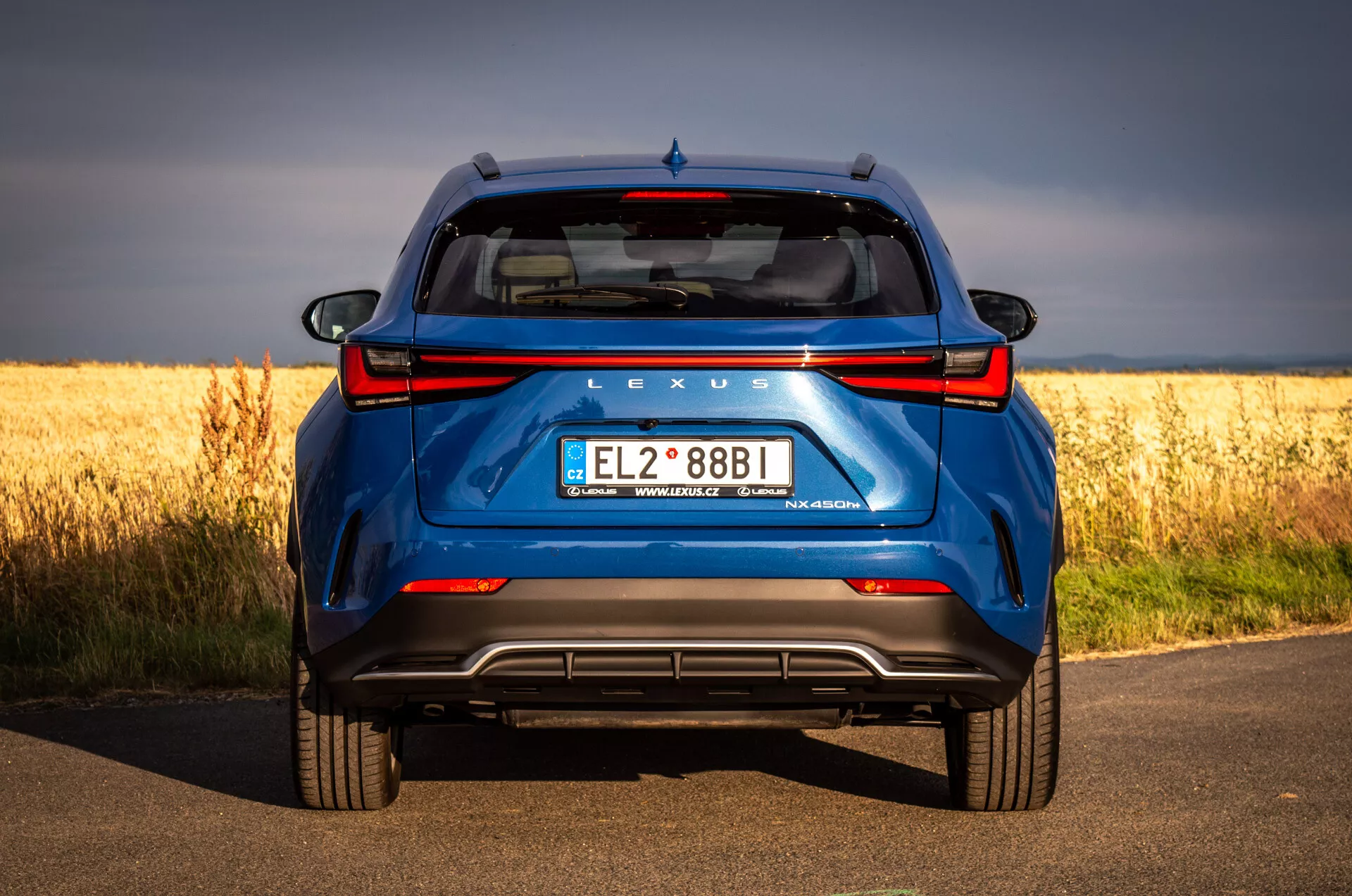 Zadní pohled na modré SUV Lexus NX 2021 na asfaltové silnici s pozadím zlatého pole, zdůrazňující hybridní technologii a designové prvky